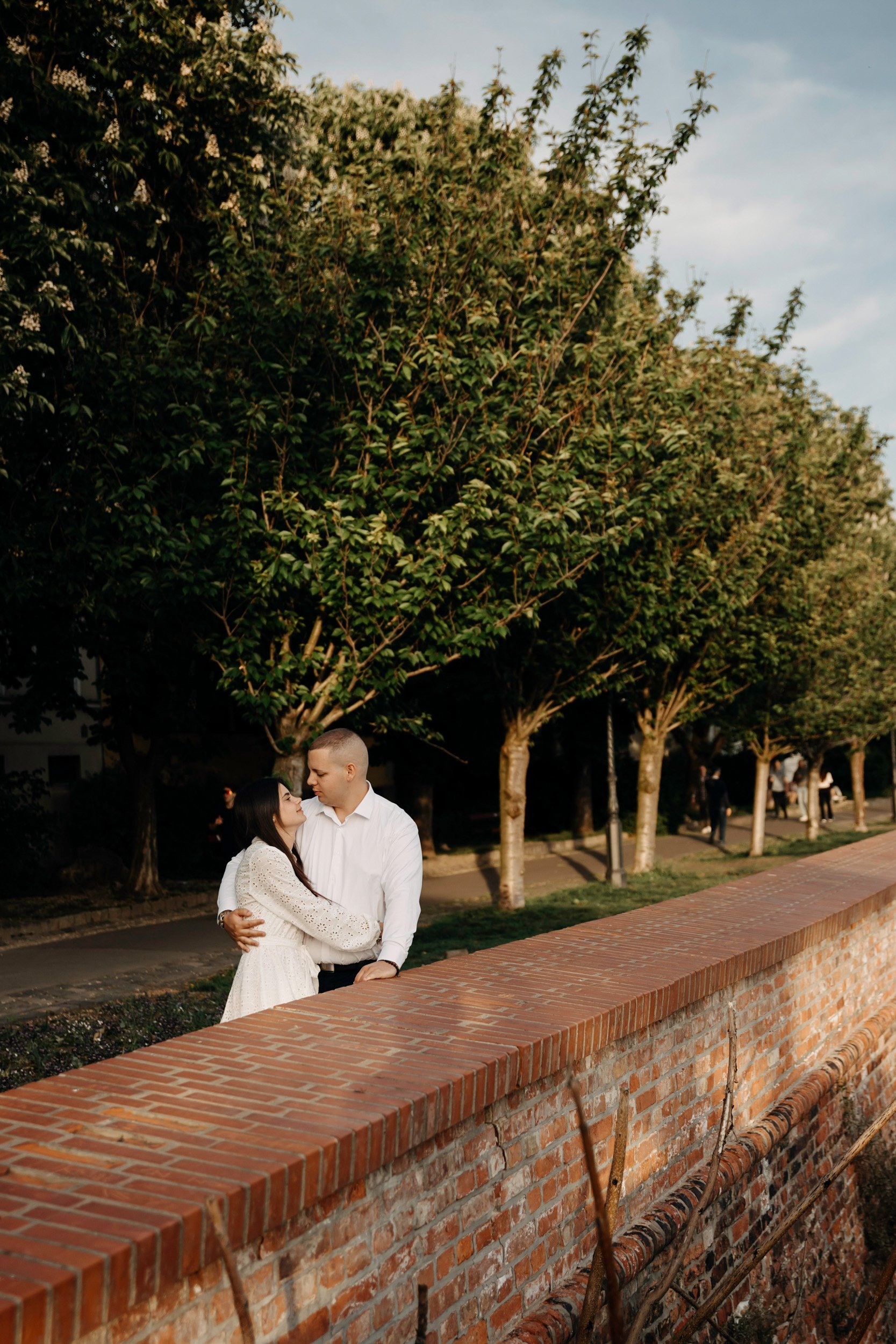 A couple stands close together outdoors near a brick wall, with a row of trees and several people walking in the background, under a partly cloudy sky.