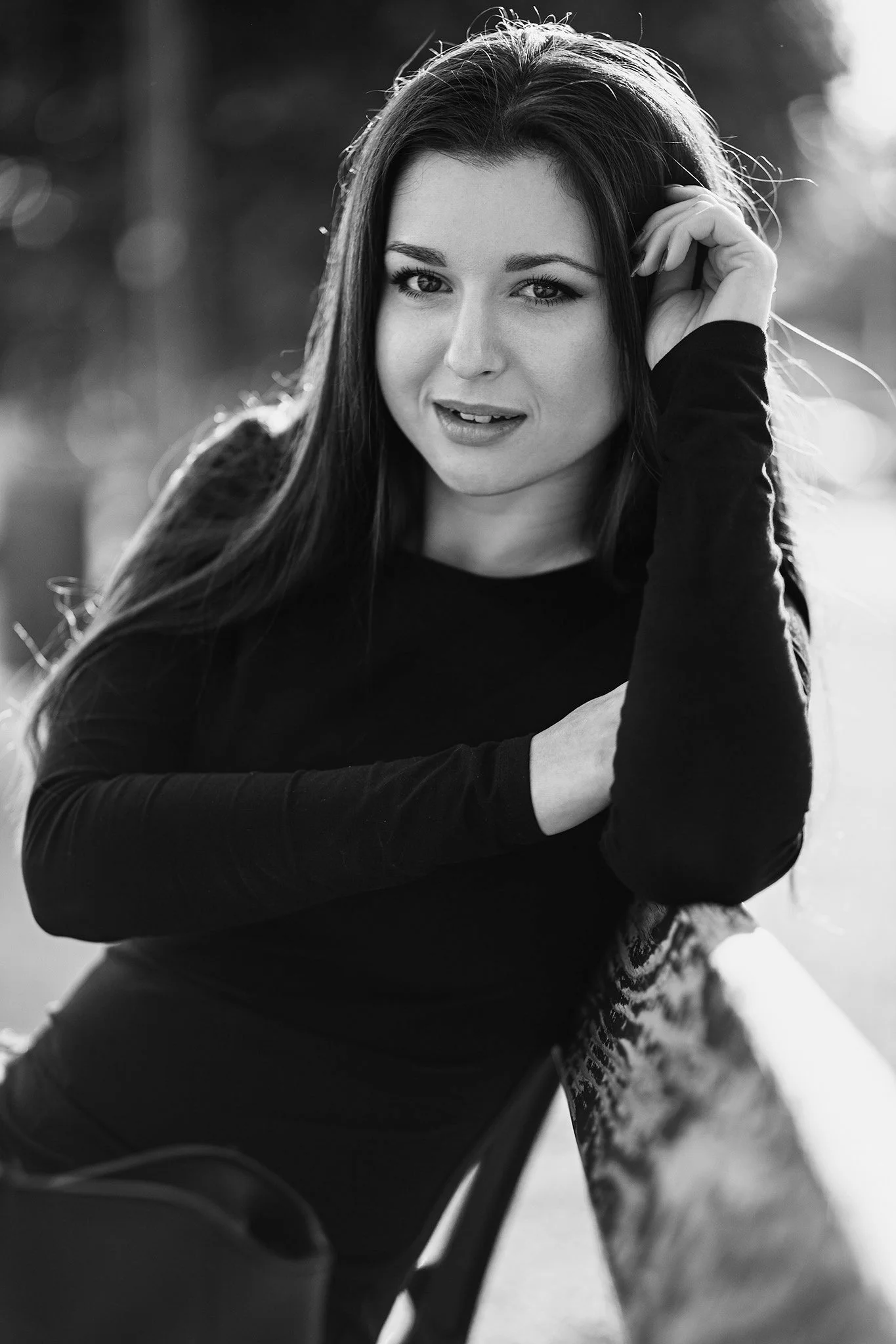 A black and white photo of a woman with long dark hair, smiling, touching her hair with her right hand, sitting outdoors with a blurred background.