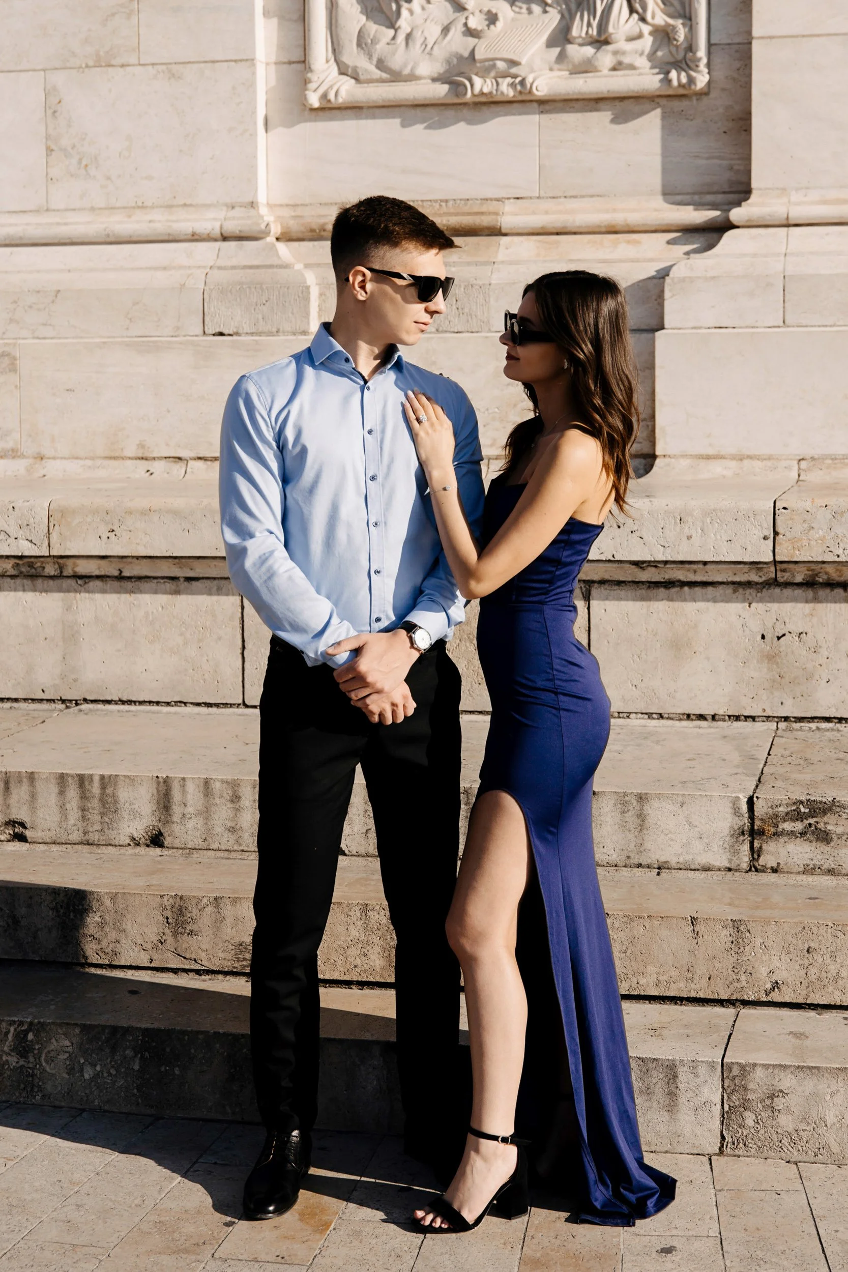 A man and a woman stand close together on stone steps in front of an ornate stone wall. They are wearing sunglasses, with the woman has long dark hair and the man has short hair. The woman is touching the man's chest with her left hand, showing a rin