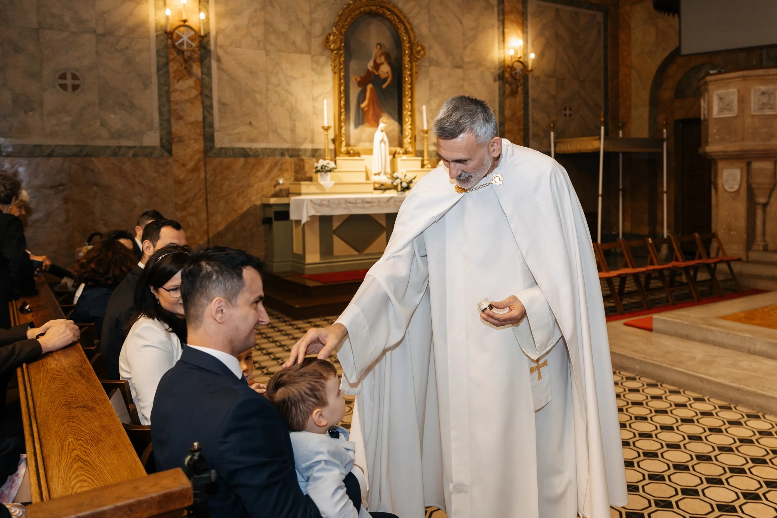 A priest in white robes with gold accents is performing a religious ceremony with children and adults seated in a church. The church’s interior features marble walls, a religious painting in a decorative gold frame, and lit candles.