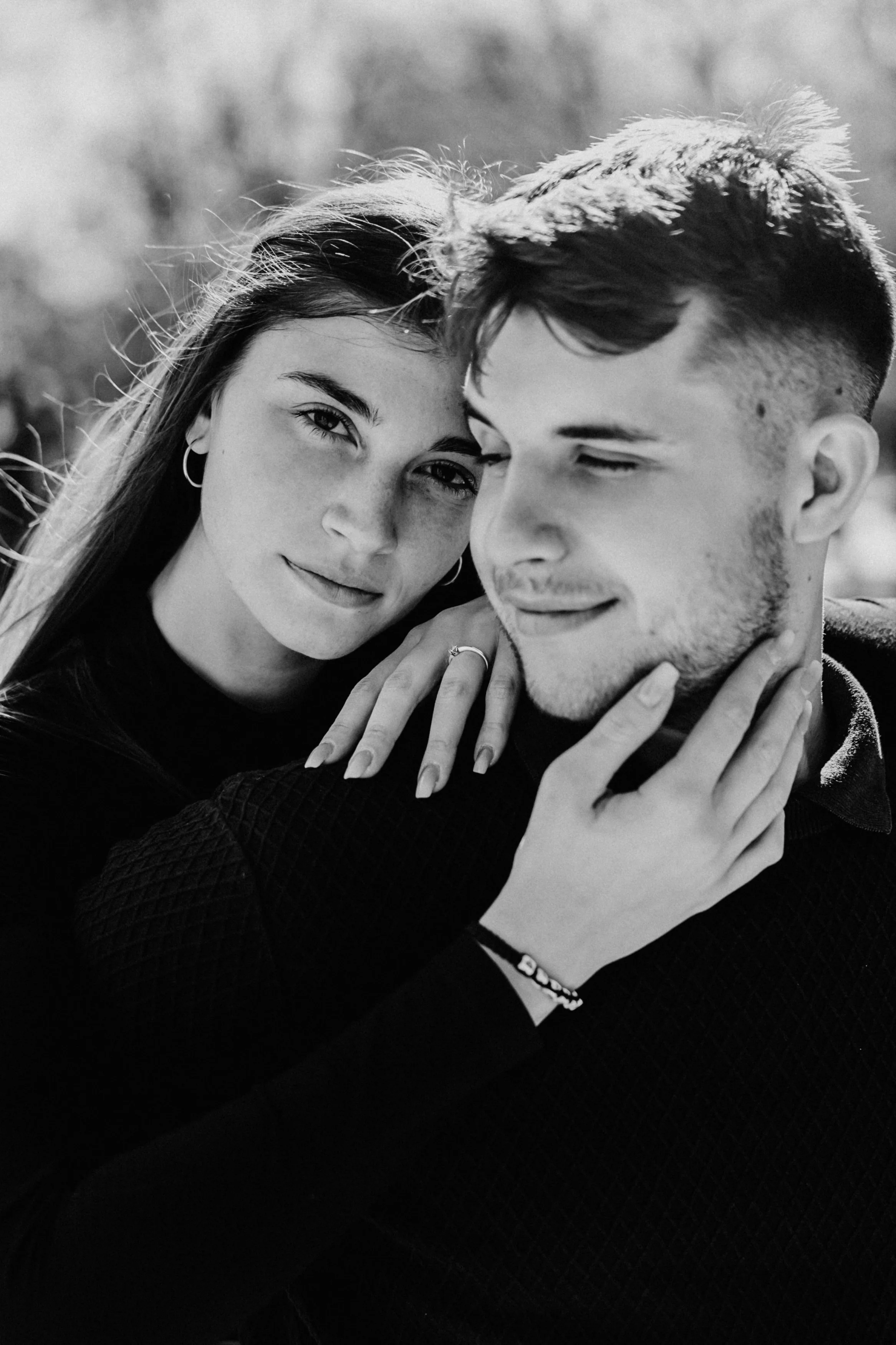 A black and white photograph of a young woman with light skin and long hair, and a young man with light skin and short hair, embracing each other outdoors. The woman has her hand on the man's neck, and they are close together with their eyes partiall