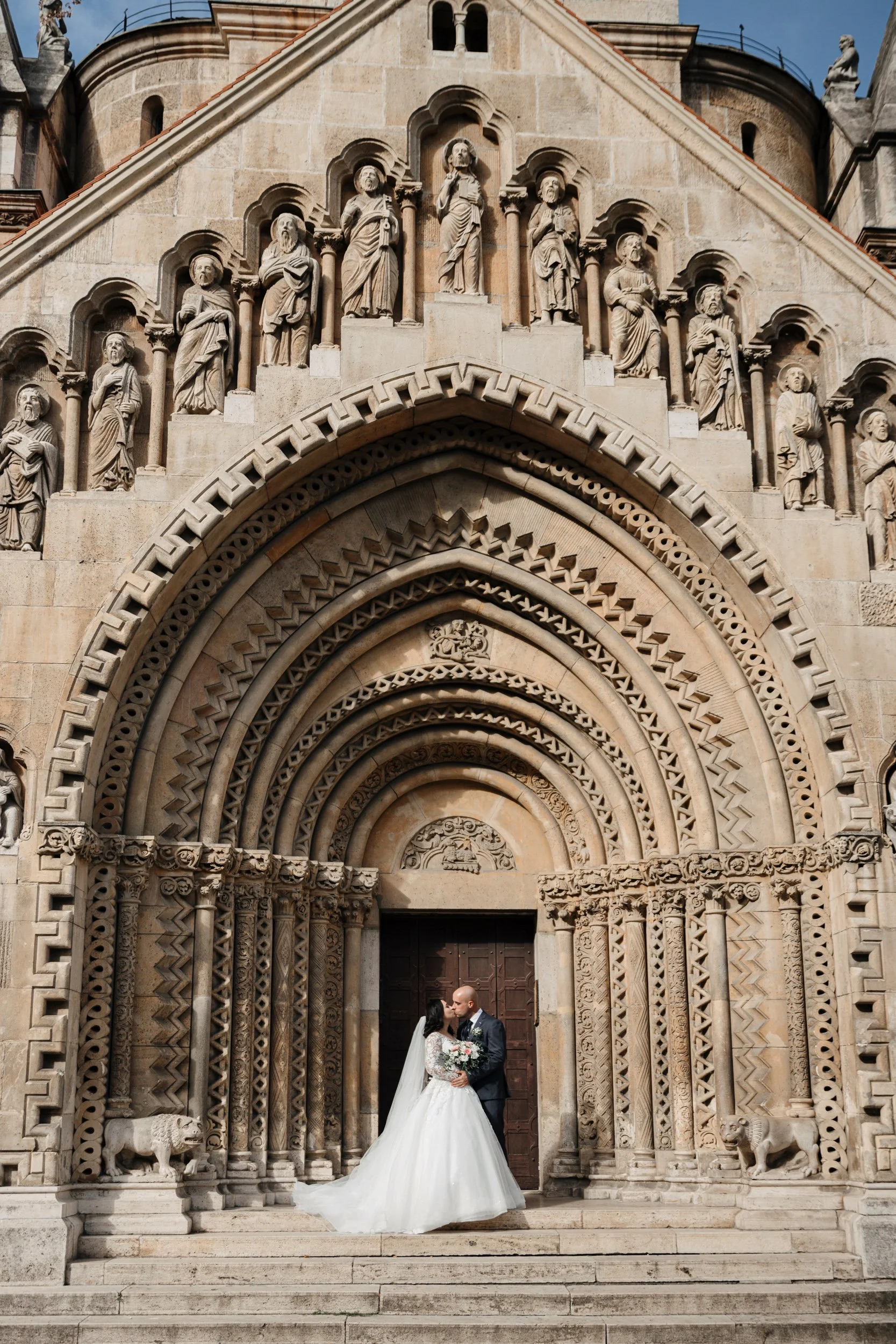 A bride and groom standing close, embracing in front of an ornate stone church entrance with intricate carvings and statues, during a wedding ceremony.