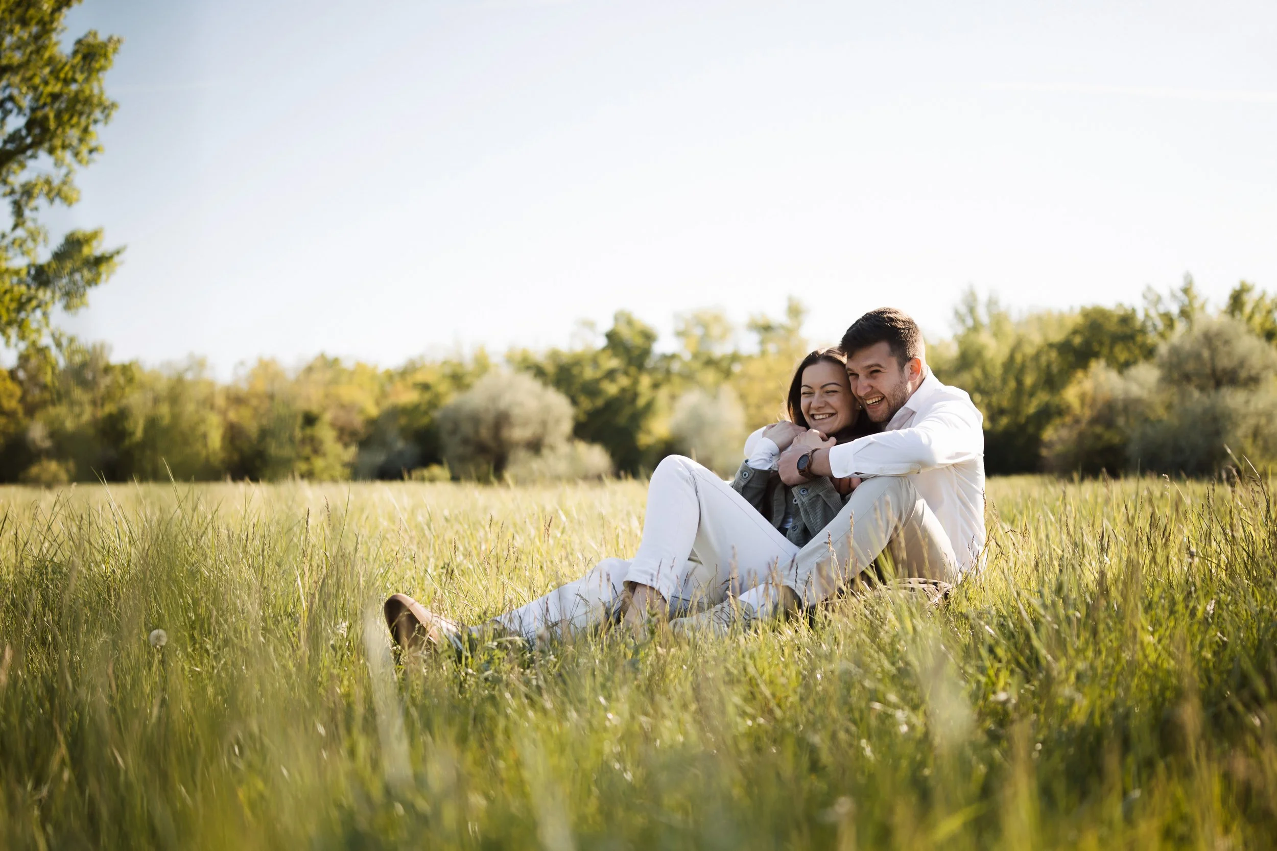 A happy couple sitting together in a grassy field on a sunny day, embracing and smiling.