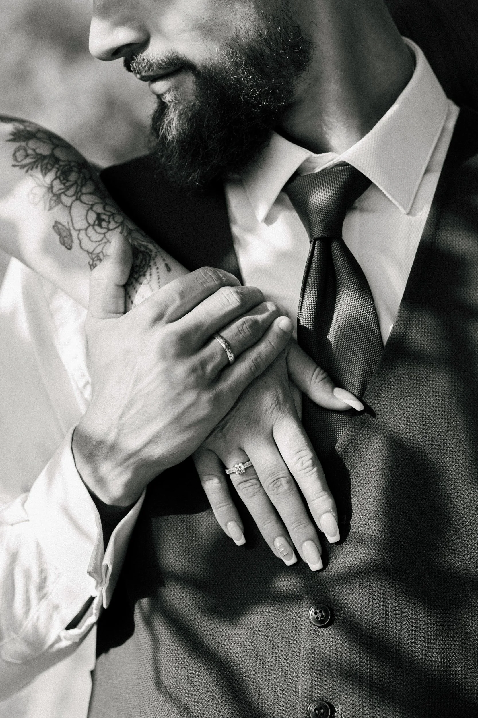 Close-up black-and-white photo of a man and woman holding hands, focusing on their hands, rings, and part of their faces and upper bodies. The woman has a floral tattoo on her arm and is wearing a ring, while the man is dressed in a shirt, tie, and v