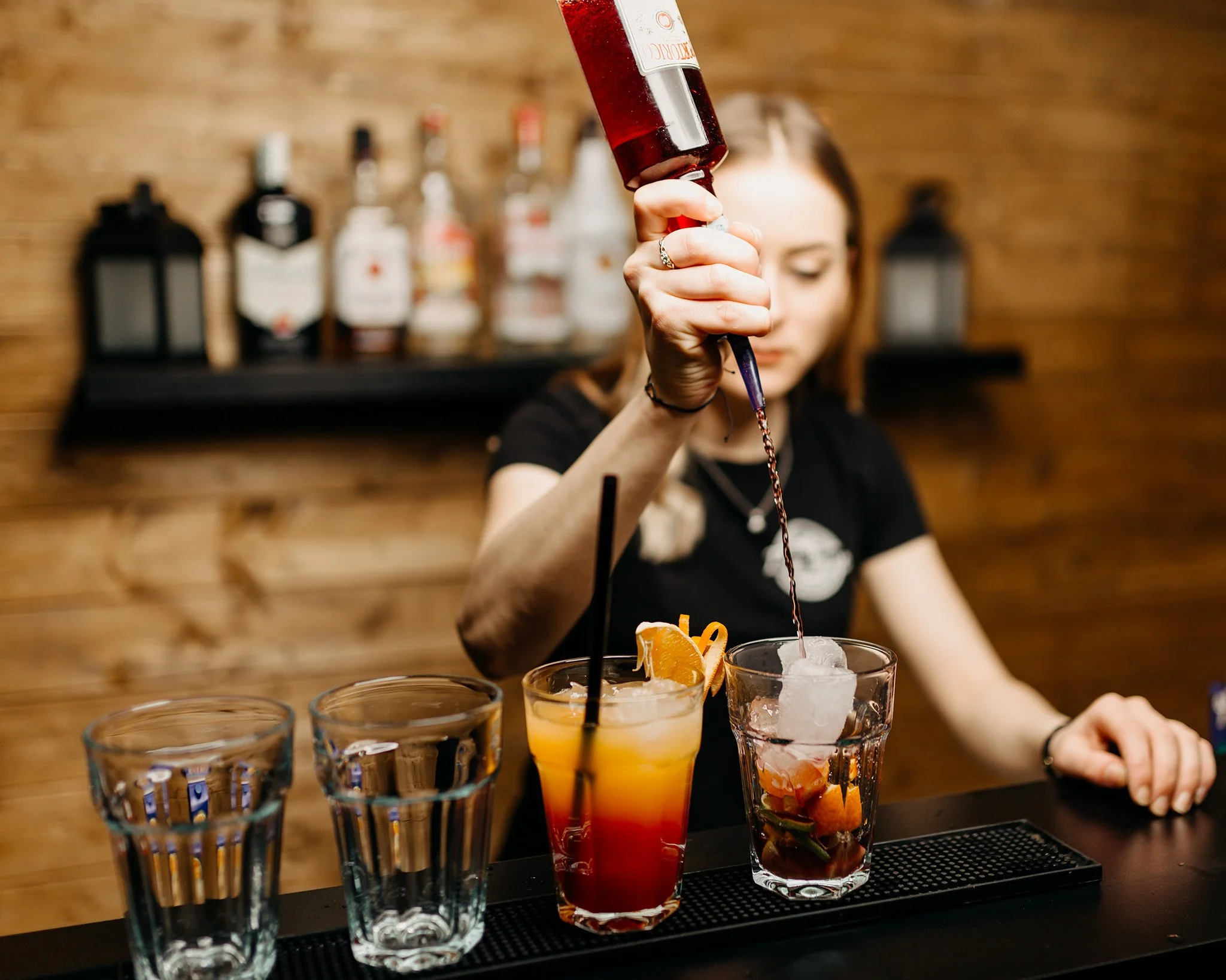 A woman in a black shirt pours a red cocktail into a decorative glass with ice and orange garnishes, surrounded by several empty glasses on a bar counter. Bottles of alcohol are visible in the background on a wooden wall.