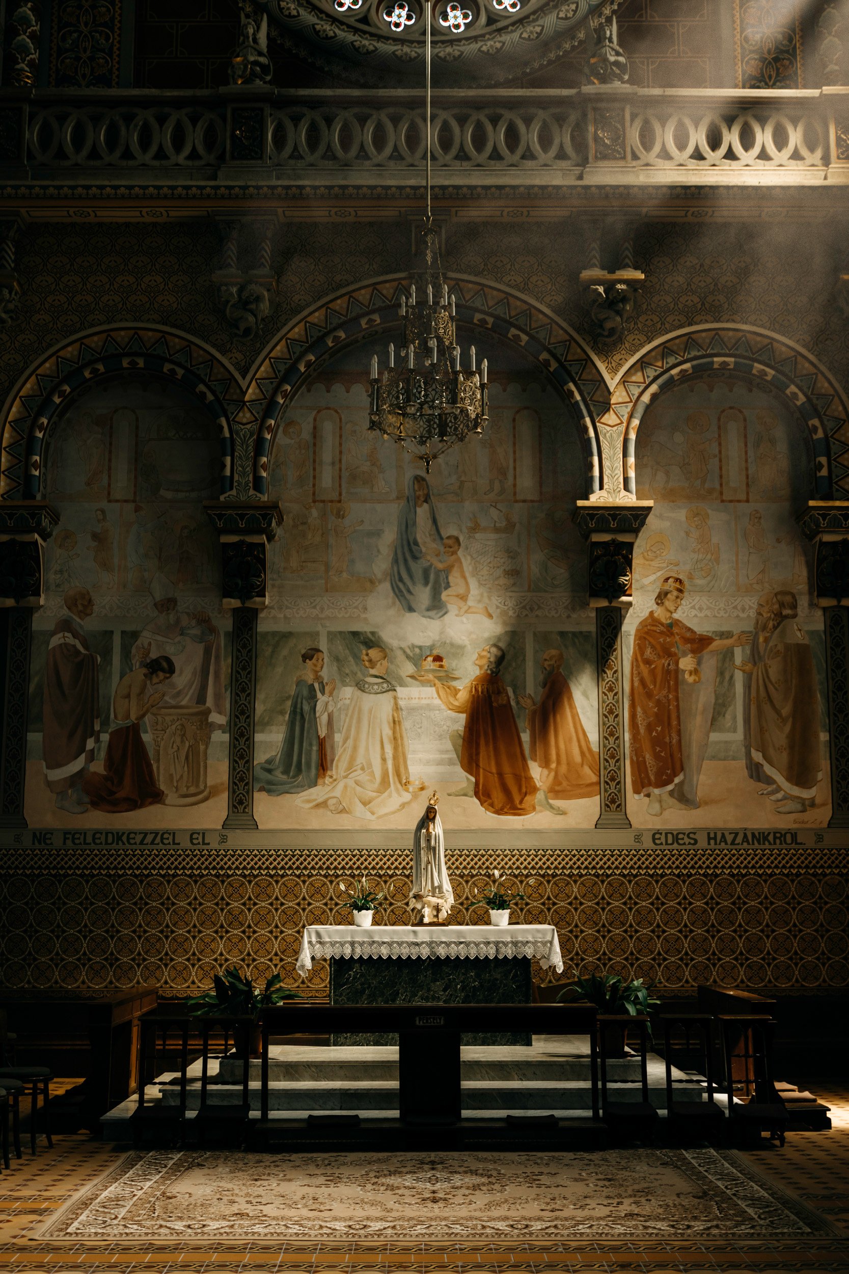 Interior of a church with an altar featuring a statue of the Virgin Mary, a white cloth, and potted plants. Behind the altar is a large mural depicting religious figures, and a chandelier hangs from the ceiling. The walls are decorated with detailed 