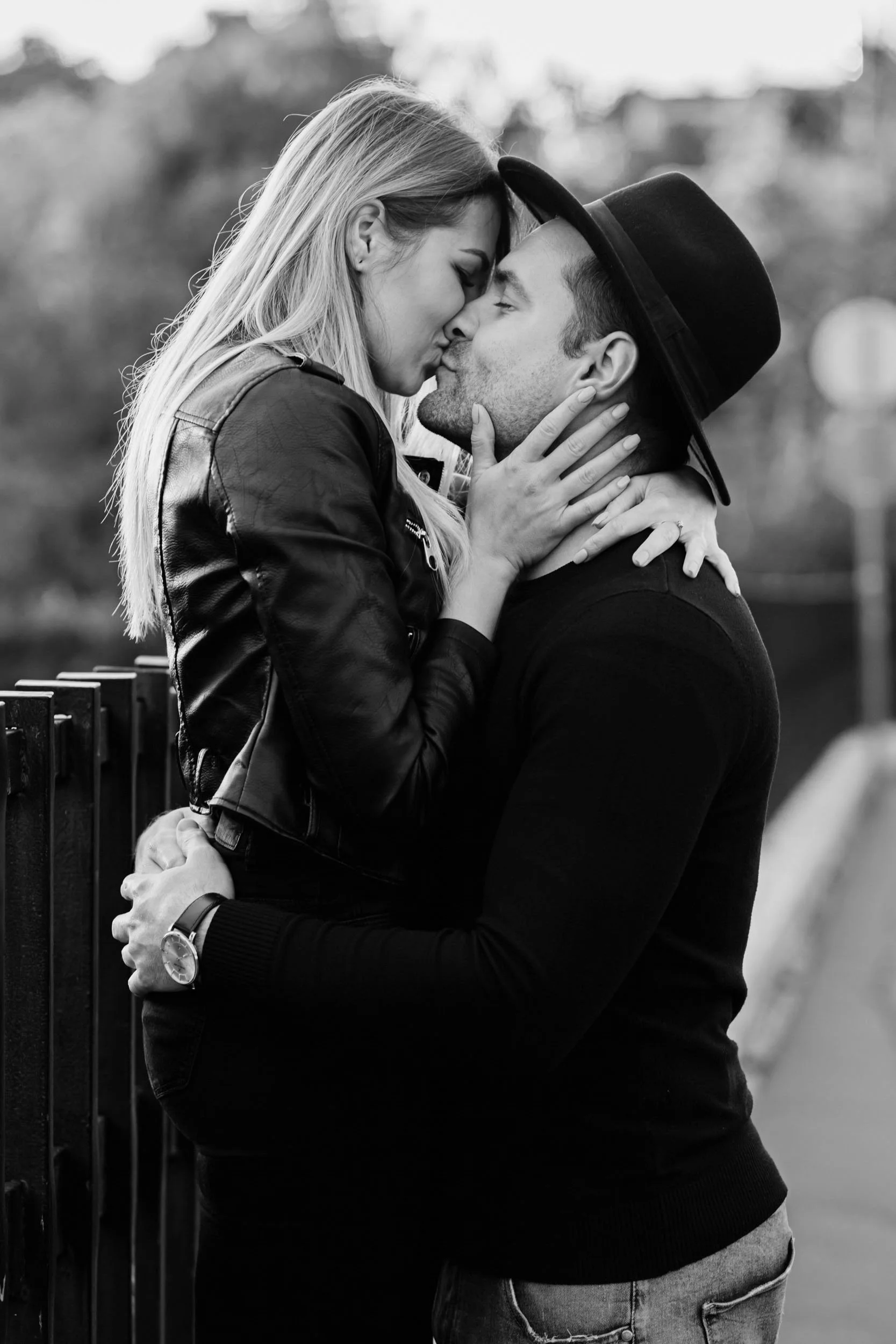 A black and white photo of a couple sharing a kiss, with the woman sitting on a bench and the man standing. The woman has long hair and is wearing a leather jacket, while the man is wearing a hat and a dark sweater.