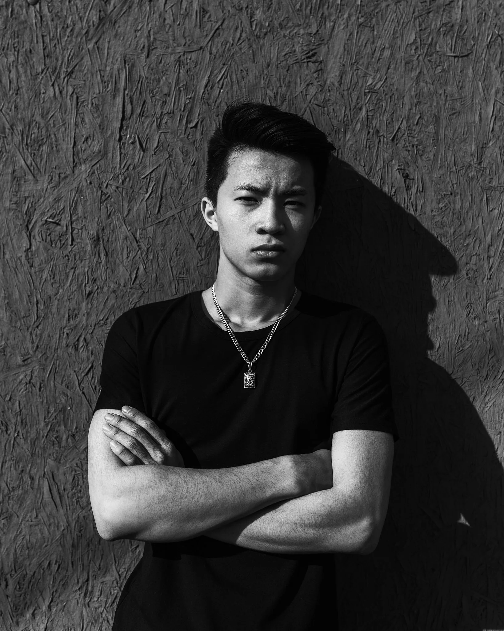Black and white photograph of a young man with short hair, wearing a chain necklace and a t-shirt, standing in front of a textured wall, crossing his arms and looking directly at the camera.