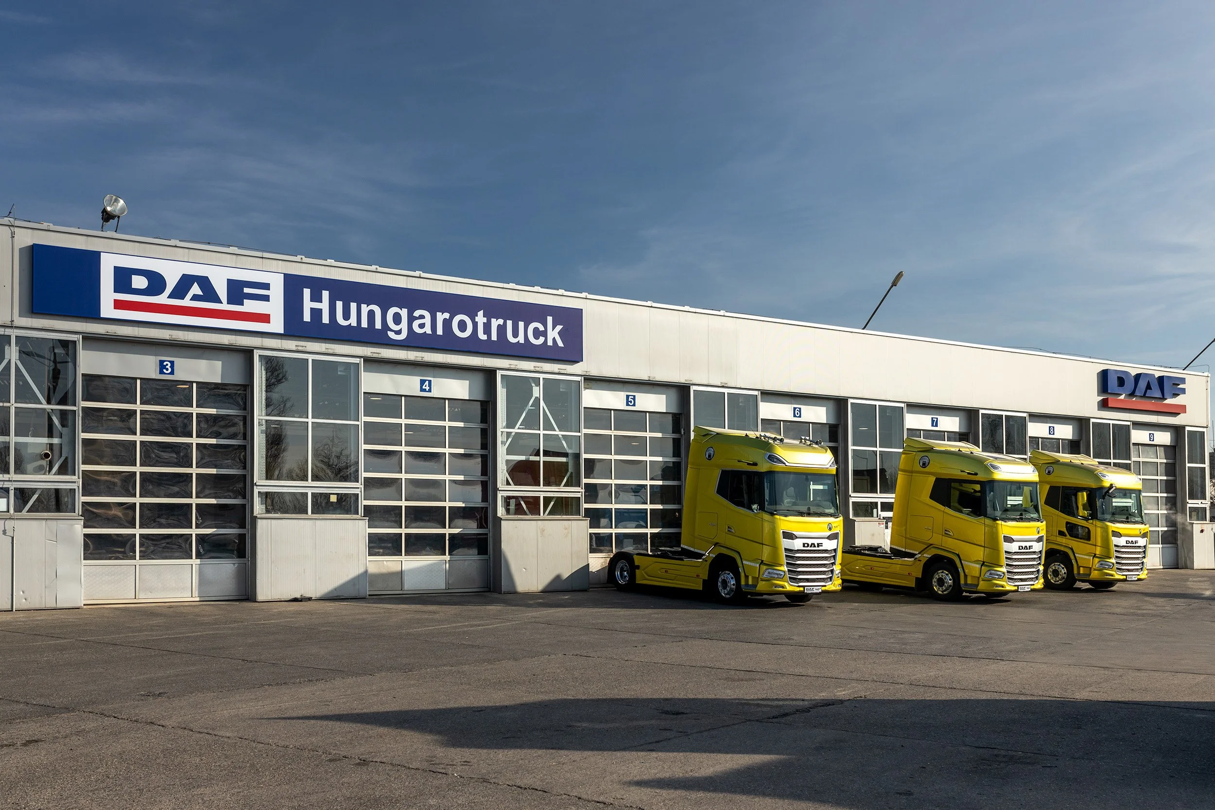 A building with signs that read 'DAF Hungarotruck' and a row of yellow trucks parked in front, under a blue sky.