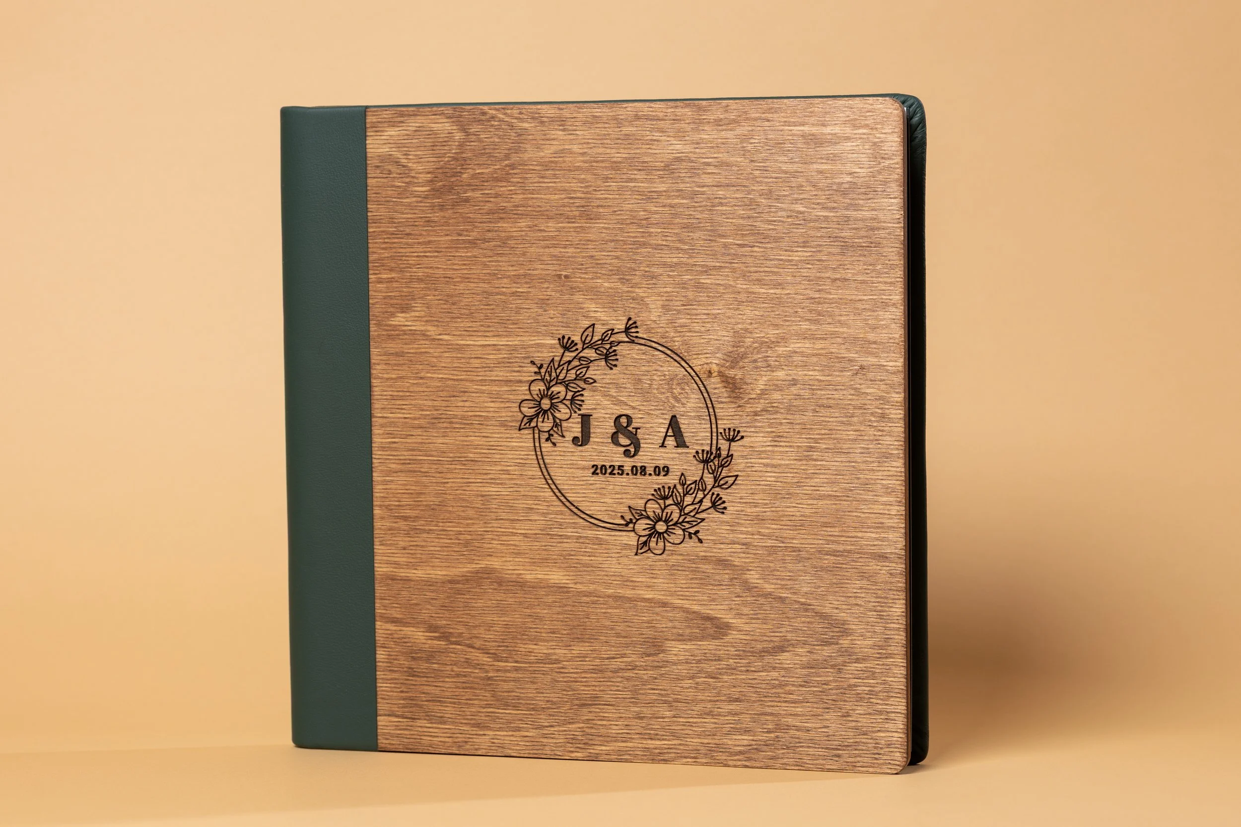 A closed hardcover book with a woodgrain pattern on the cover, featuring a decorative floral circle and the initials J & A, along with a date 2025.08.09. The spine and edges are dark green, and the background is a plain beige surface.