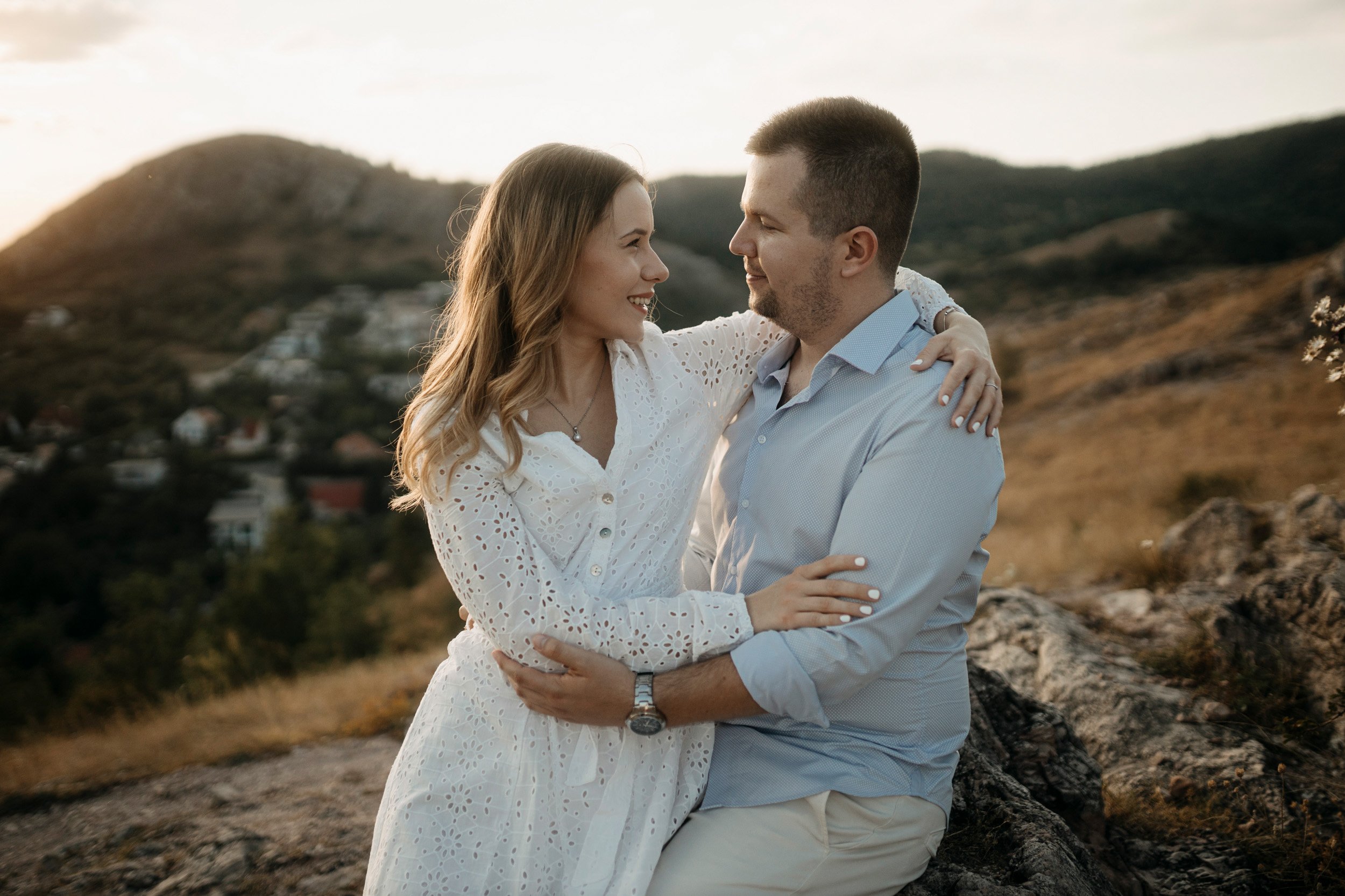 A couple embracing outdoors, looking into each other's eyes against a mountainous landscape at sunset.