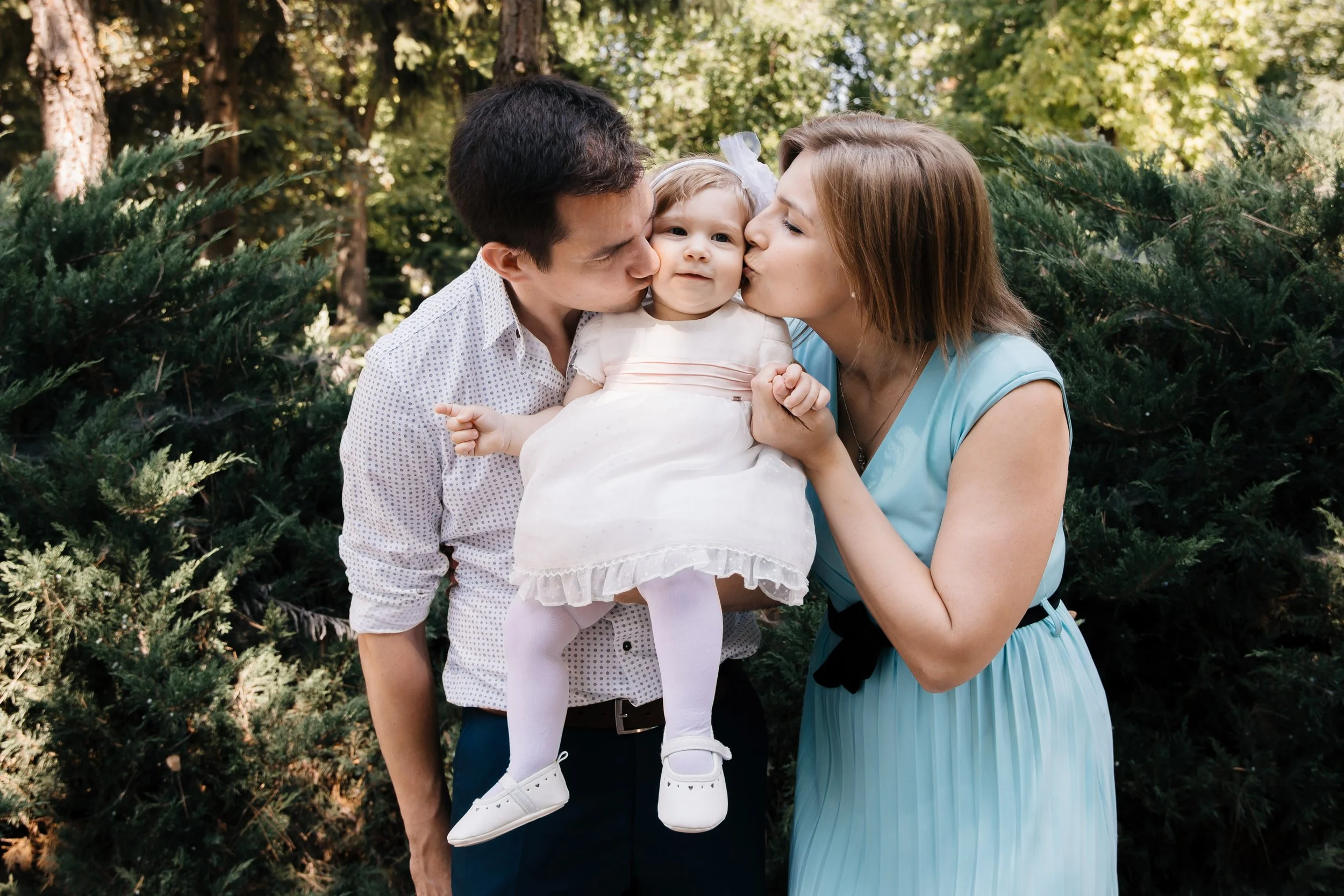 A family of three standing outdoors among greenery, with the father and mother kissing their young daughter on the cheeks.