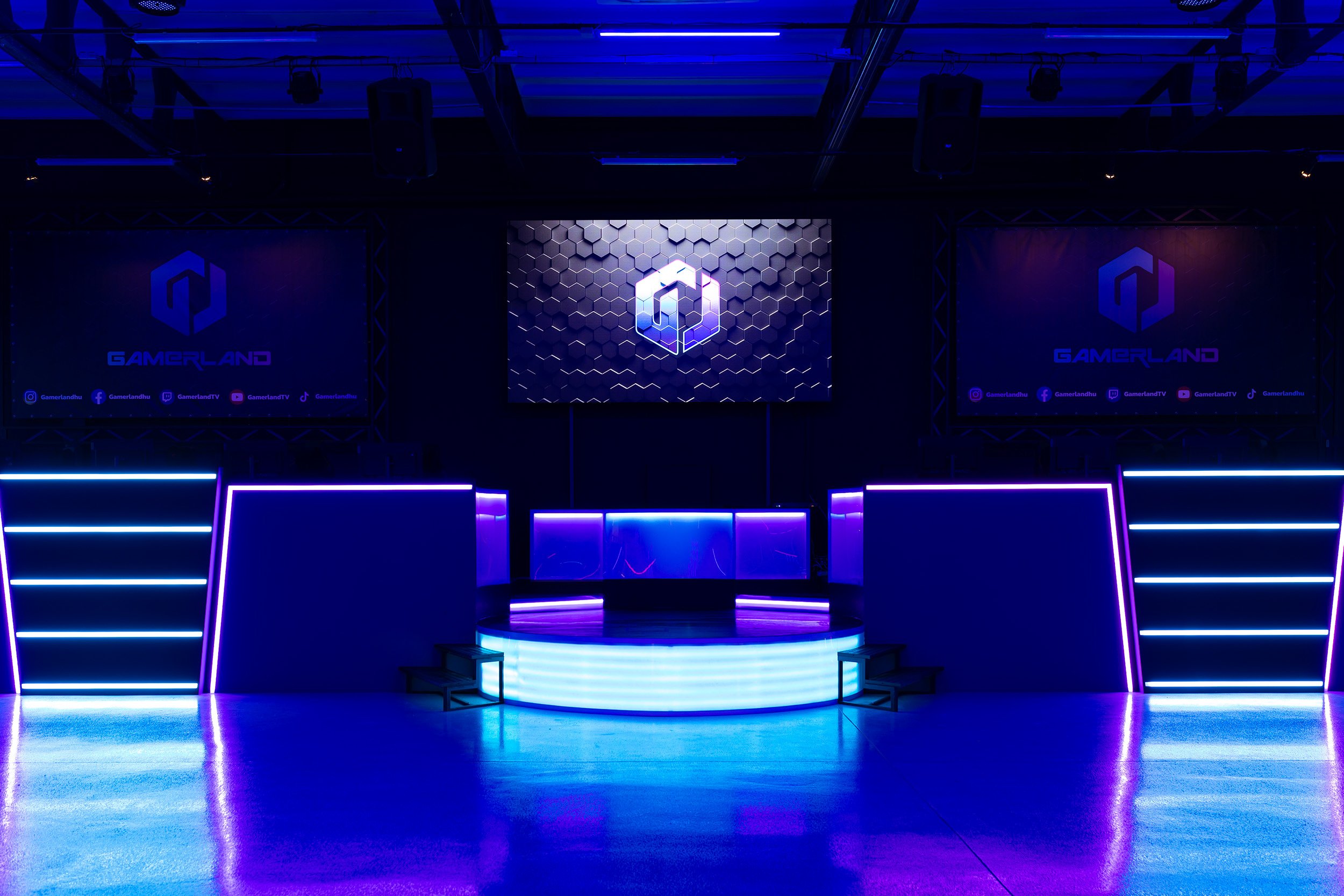 Gamerland gaming event stage with large screens, purple and blue neon lighting, hexagonal logo, social media icons at the bottom