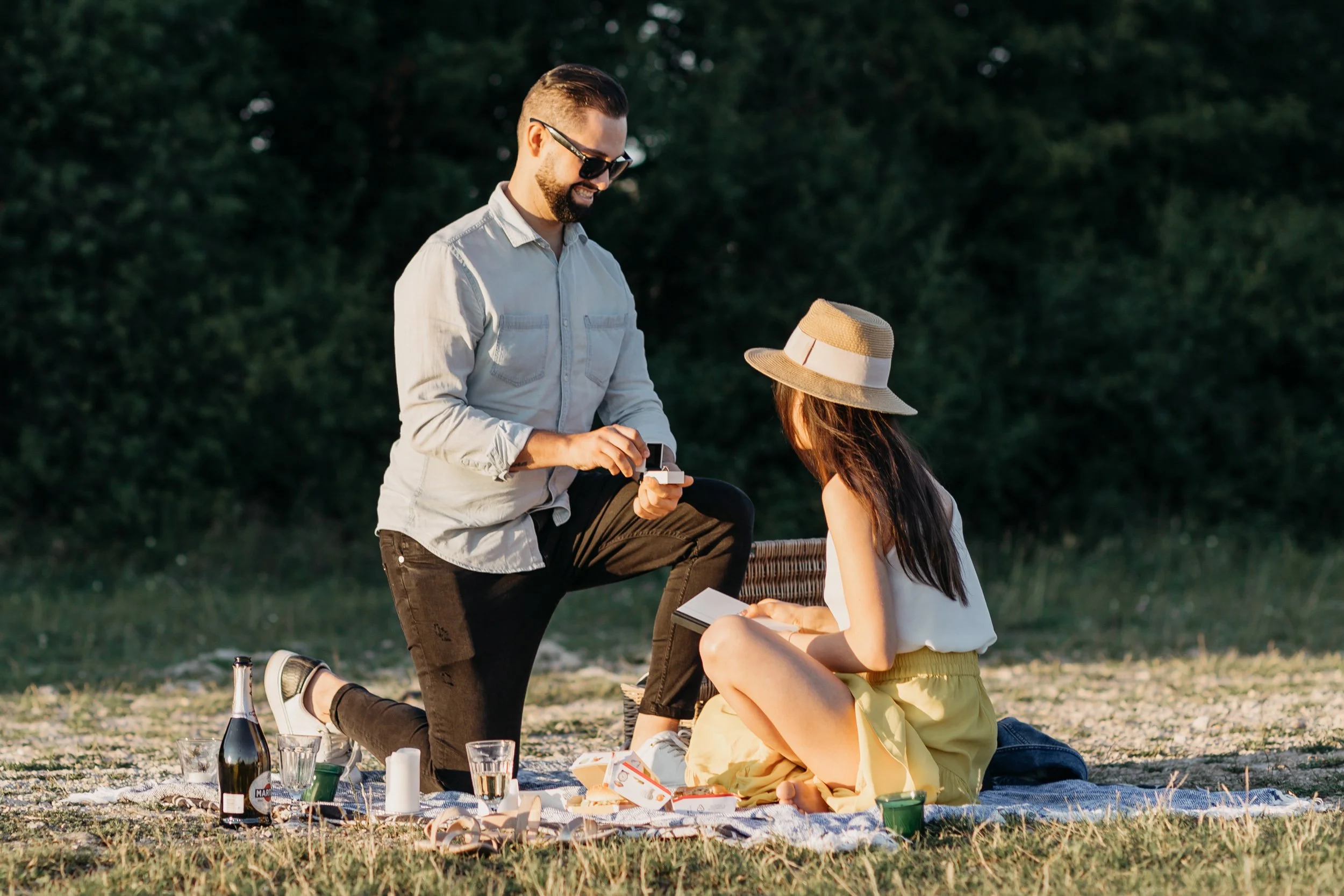 A man kneeling and a woman sitting on a blanket outdoors, exchanging a small gift, with wine bottles and glasses nearby, during a picnic in the evening.