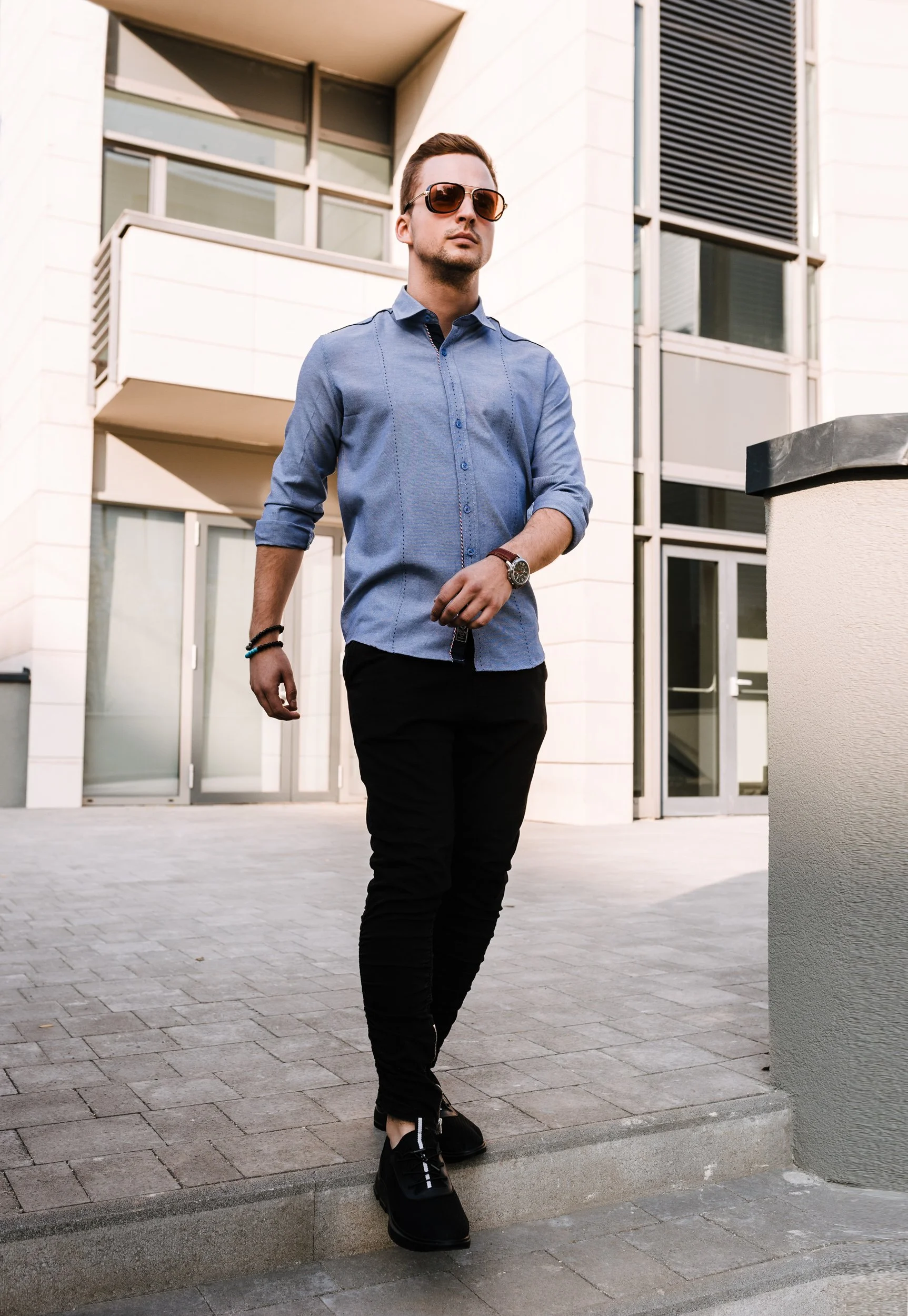 A young man walking outdoors in front of a modern building with large windows and white walls. He is wearing sunglasses, a blue button-up shirt with rolled-up sleeves, black pants, and black shoes. He is accessorized with a wristwatch, bracelets, and