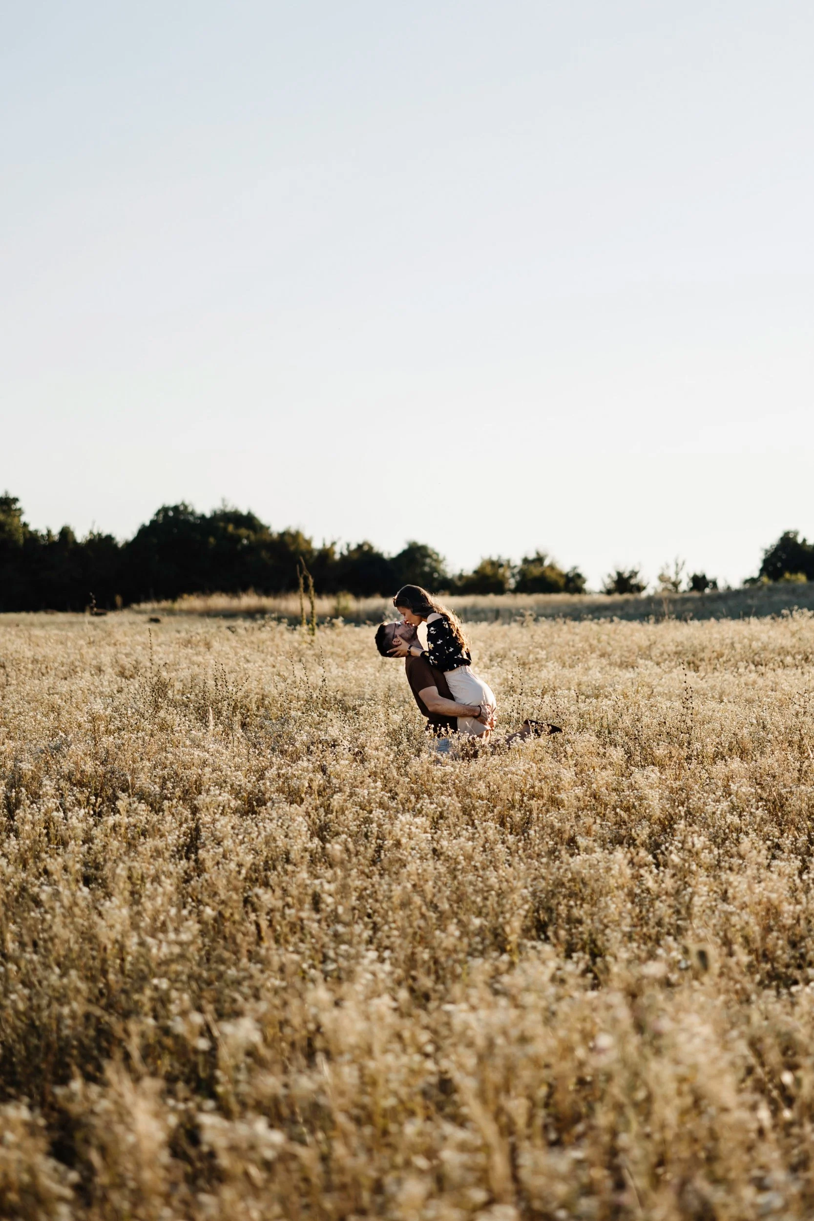 A couple kissing in a field of tall, golden grass with trees in the background under a clear sky.