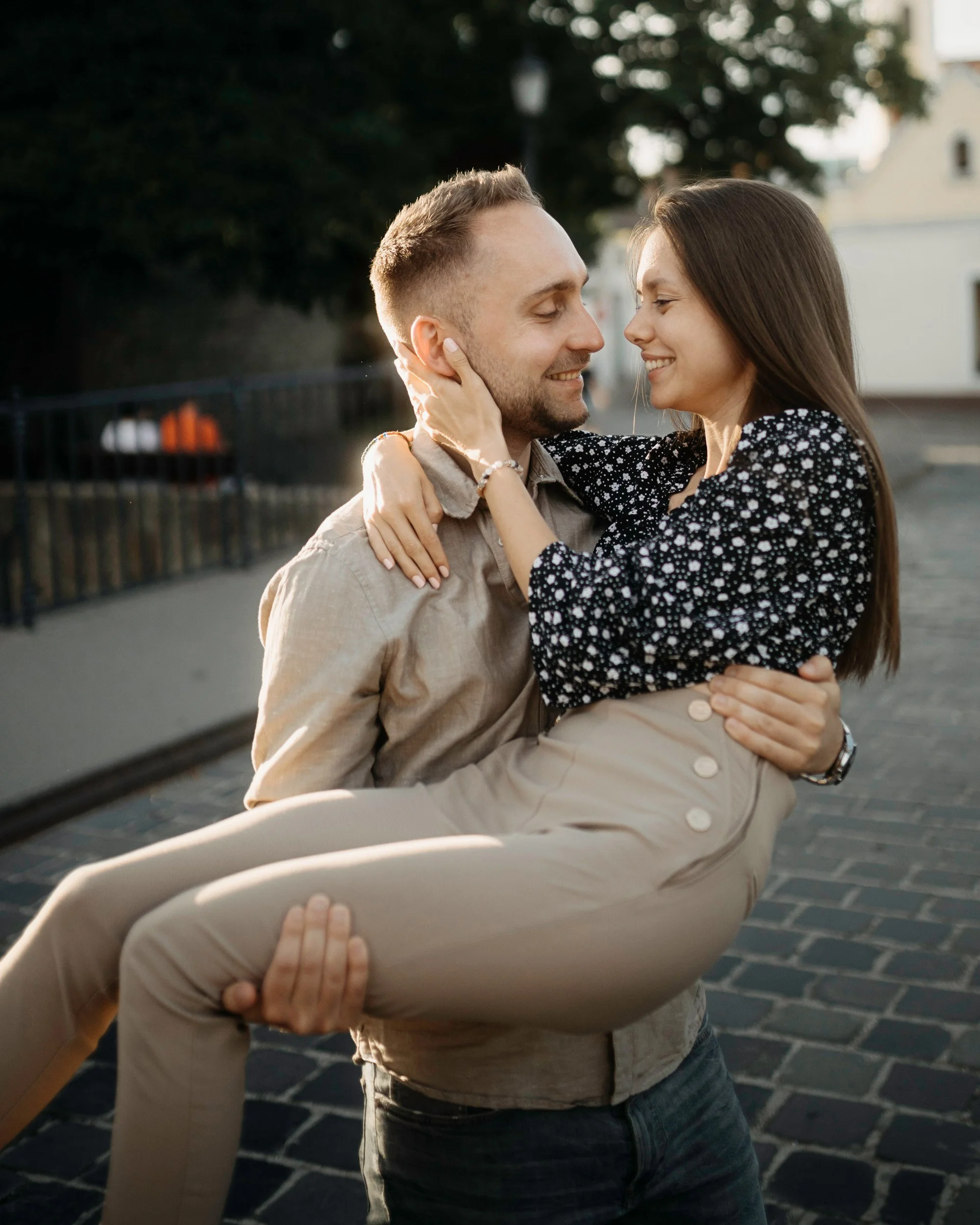 A couple hugging outdoors, with the woman holding the man's face and the man smiling with eyes closed, standing on a brick-paved street with trees and a building in the background.