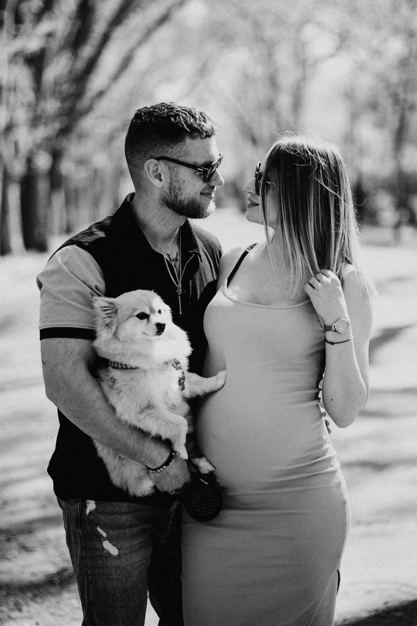 A couple standing outdoors, sharing a moment of affection, with the man holding a fluffy puppy. The woman is pregnant, and they are both wearing sunglasses, smiling and looking at each other.