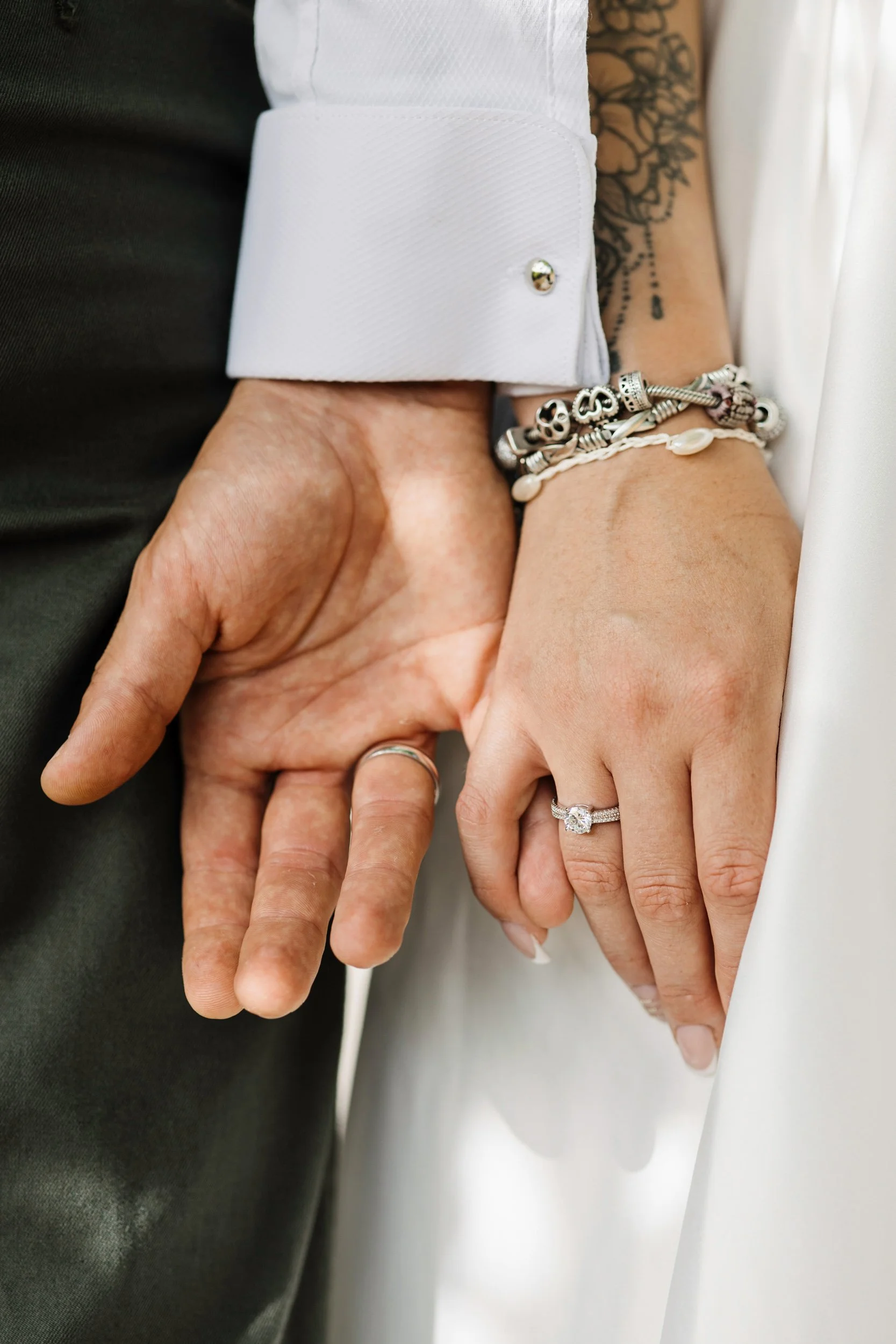 Close-up of a couple holding hands, showing wedding rings and jewelry, with the man in a dark suit and the woman in white attire, showing a tattooed arm.