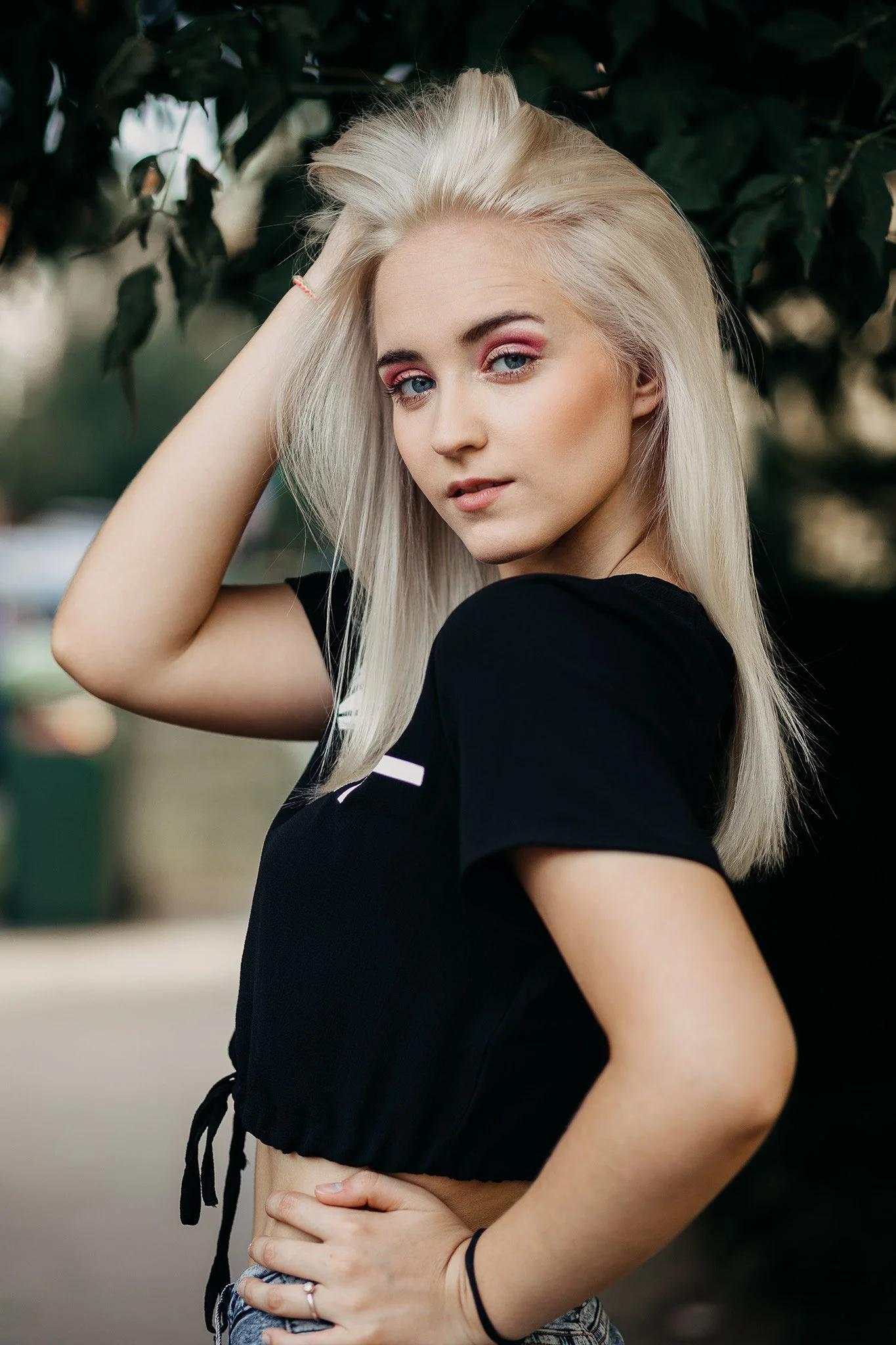 A young woman with long platinum blonde hair posing outdoors in front of green foliage. She is wearing a black crop top and denim shorts, with her left arm resting on her hip and her right hand touching her hair.