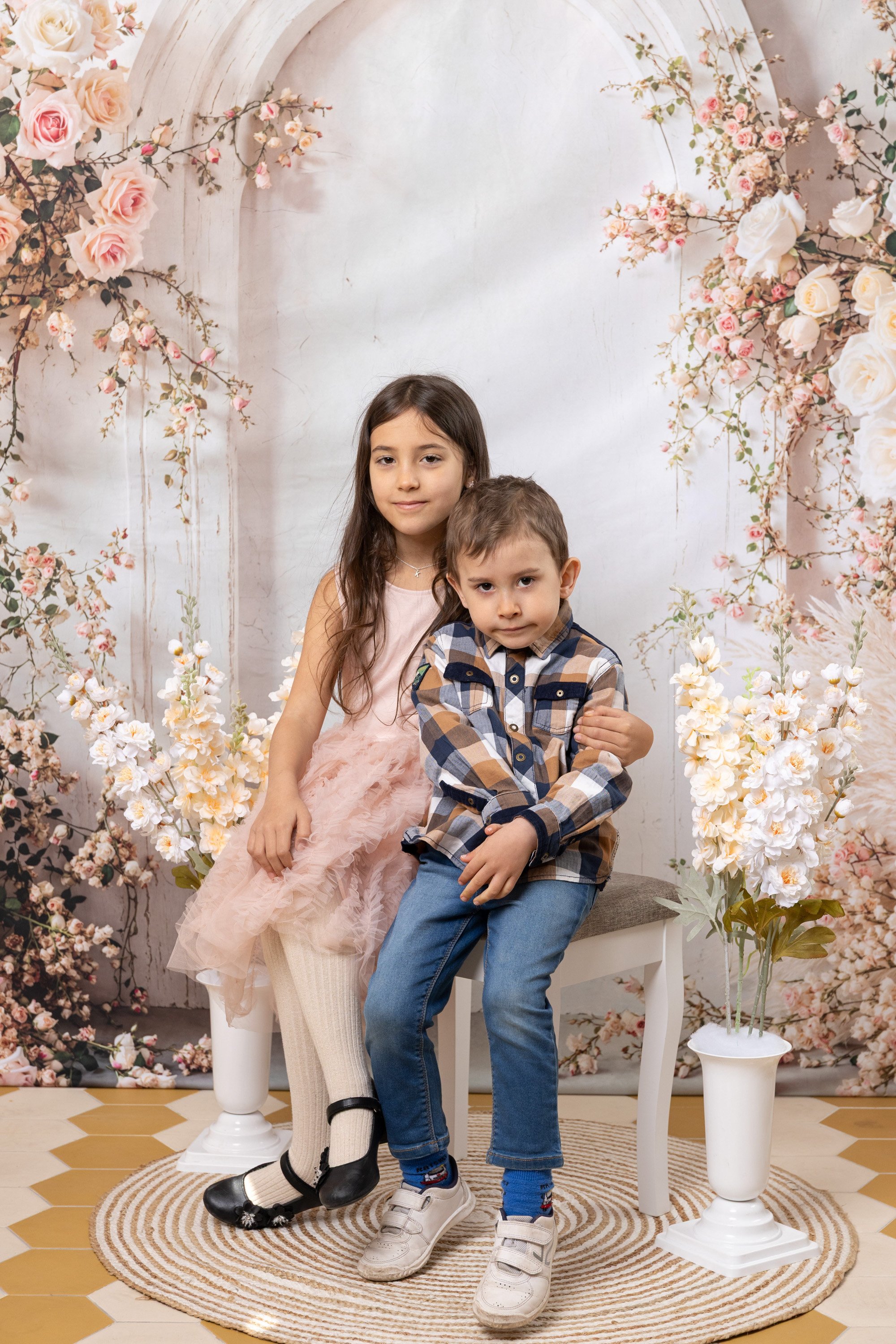 Two children, a girl in a pink dress and a boy in a plaid shirt, sitting on a white bench surrounded by pink and white flowers on a floral backdrop.