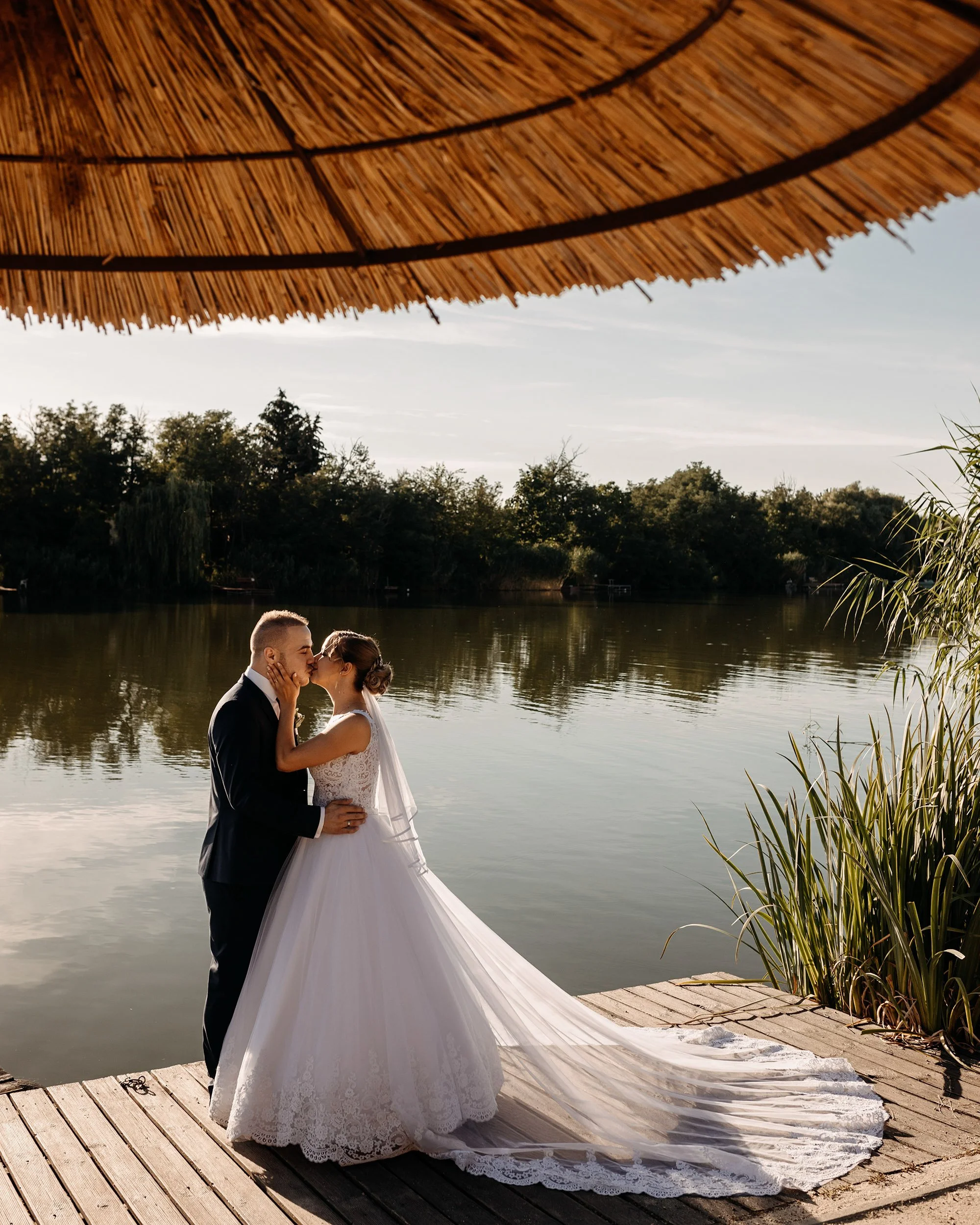 A couple, dressed in wedding attire, sharing a kiss by a lake under a large straw umbrella, with trees and cloudy sky in the background.