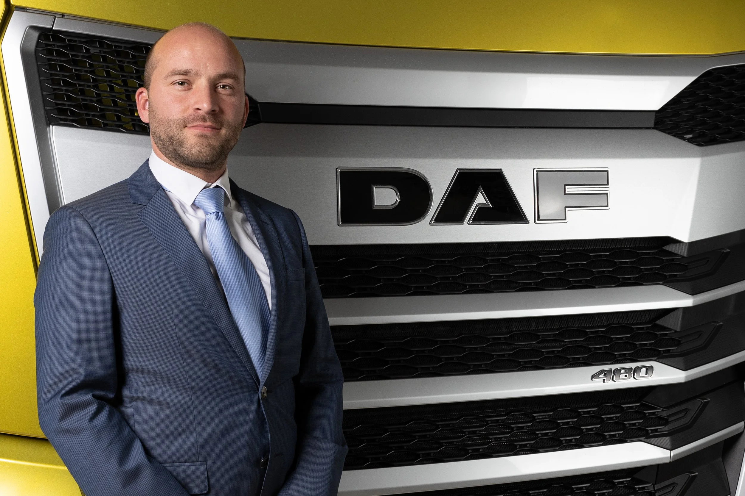 A man in a blue business suit and light blue tie standing in front of a large truck with a yellow cab and black grille, with the brand logo and model number visible.