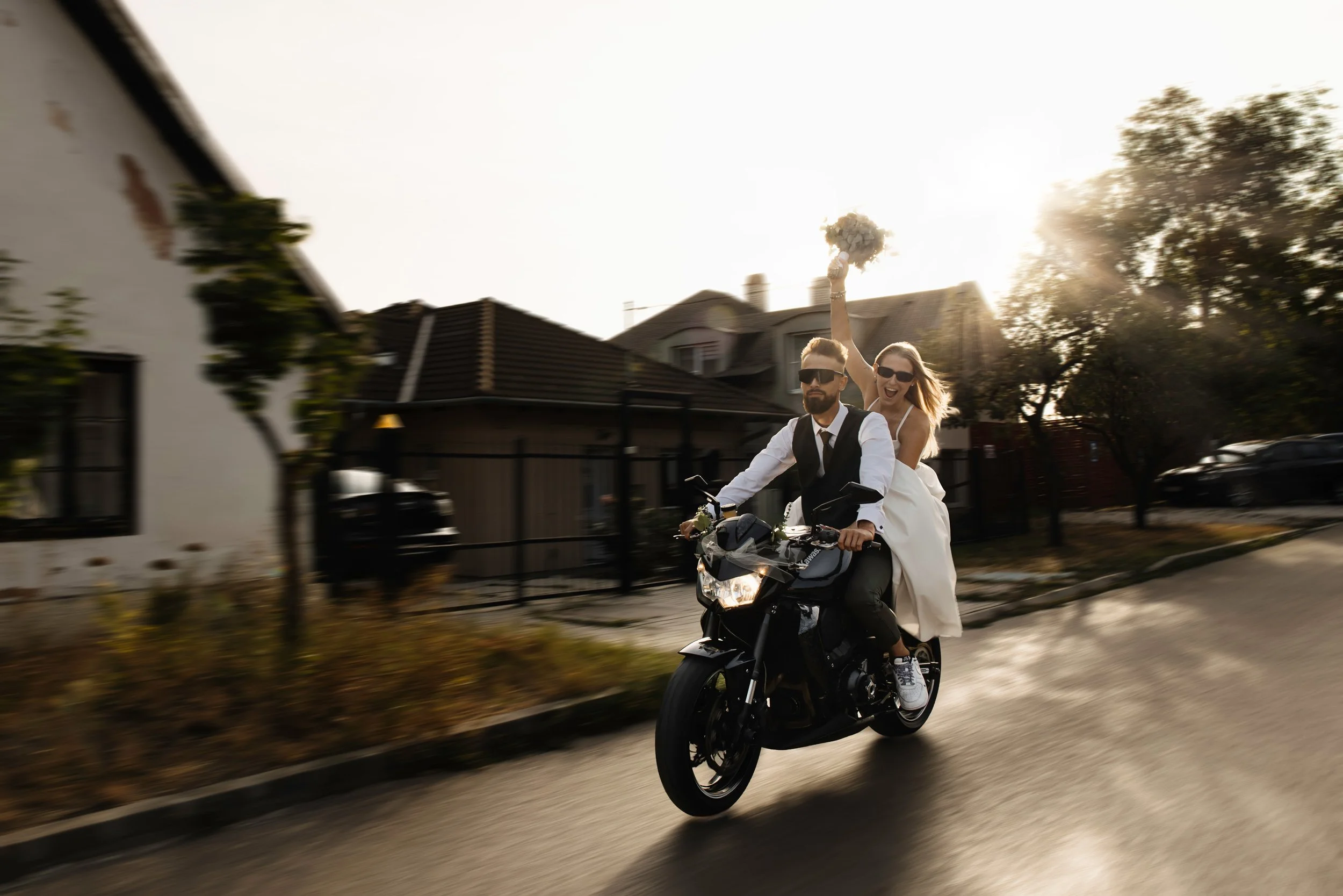 A couple riding a motorcycle down a residential street. The woman is dressed as a bride with a white dress and veil, holding a bouquet, excited and smiling, while the man is wearing sunglasses and a white shirt with a black vest, focused on riding.