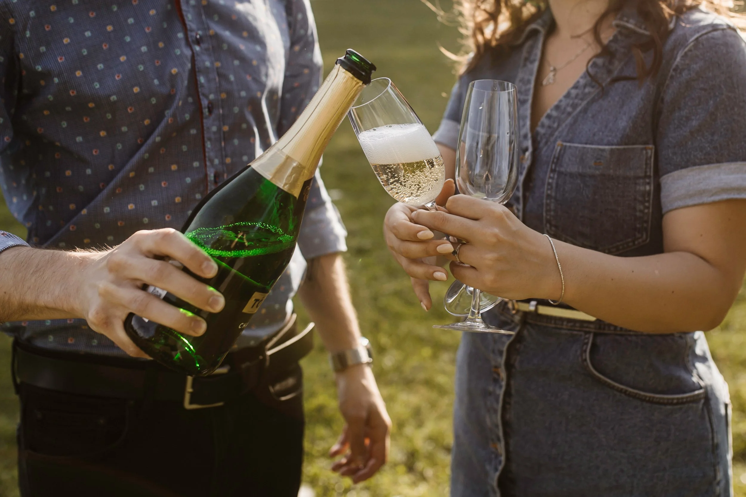Two people are holding glasses and a bottle of champagne outdoors, with one person pouring champagne into a glass. The scene is set during the daytime.