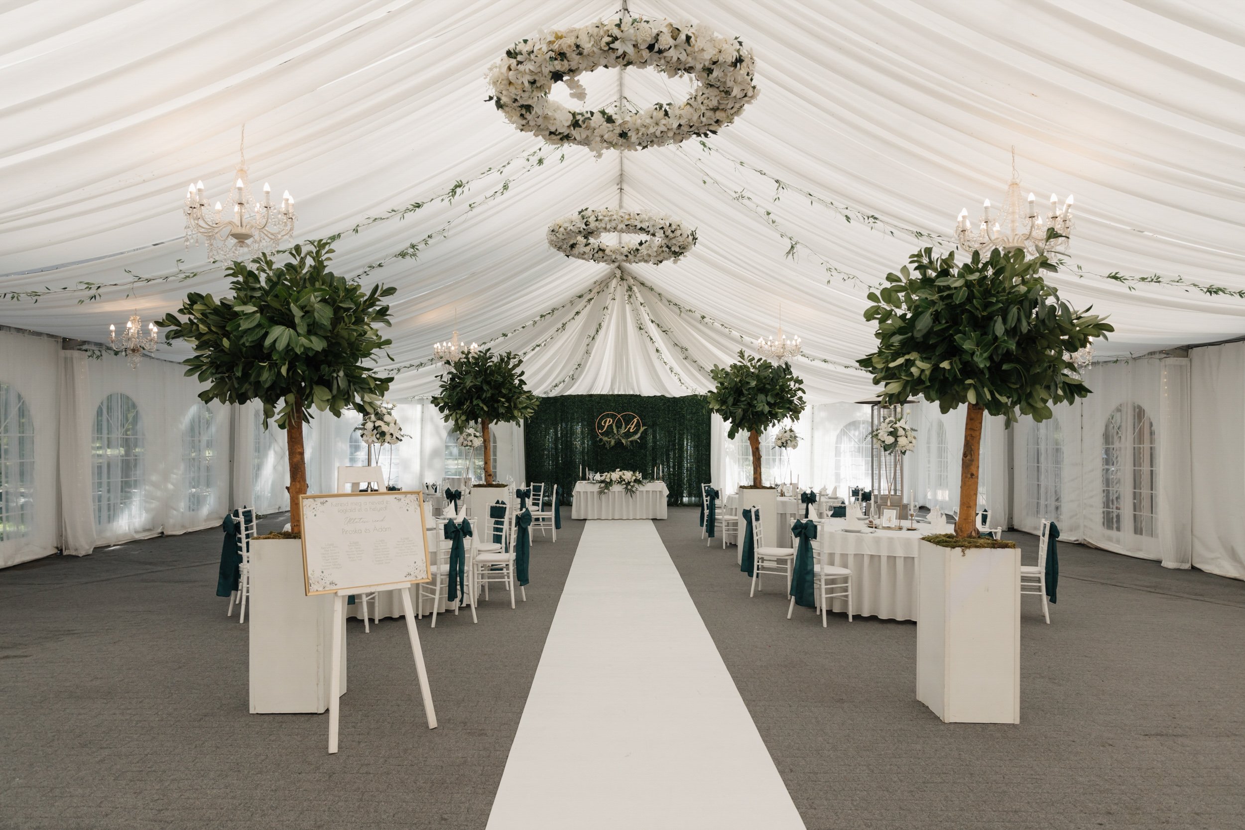 Elegant wedding reception tent decorated with hanging white floral chandeliers, green foliage, and white drapes. Central aisle lined with white chairs tied with navy bows, leading to a decorated altar with initials.