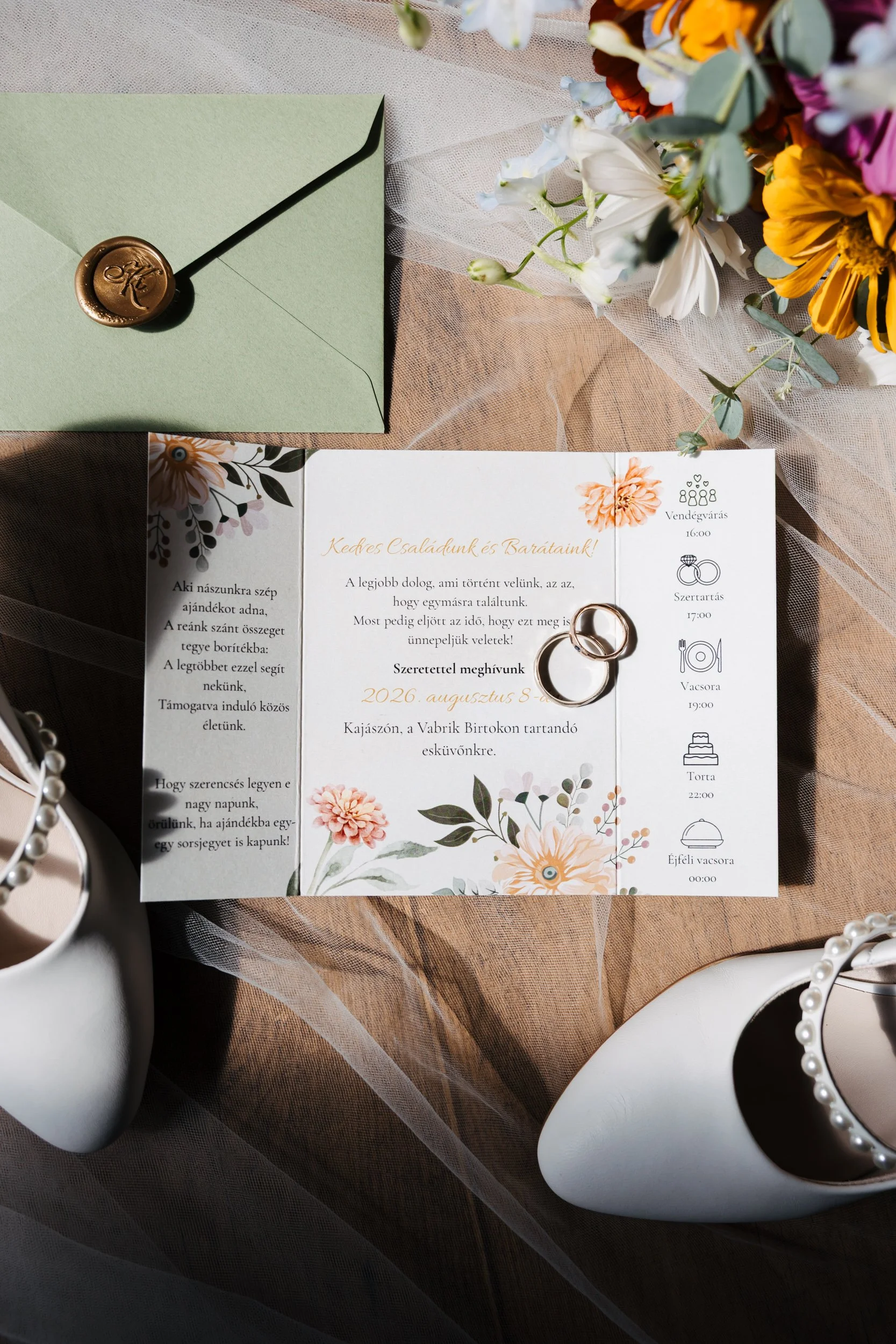 Wedding invitation card featuring floral designs, a green envelope sealed with a wax stamp, wedding rings, and surrounded by white vases and pearl necklaces on a table with a beige and sheer fabric overlay.