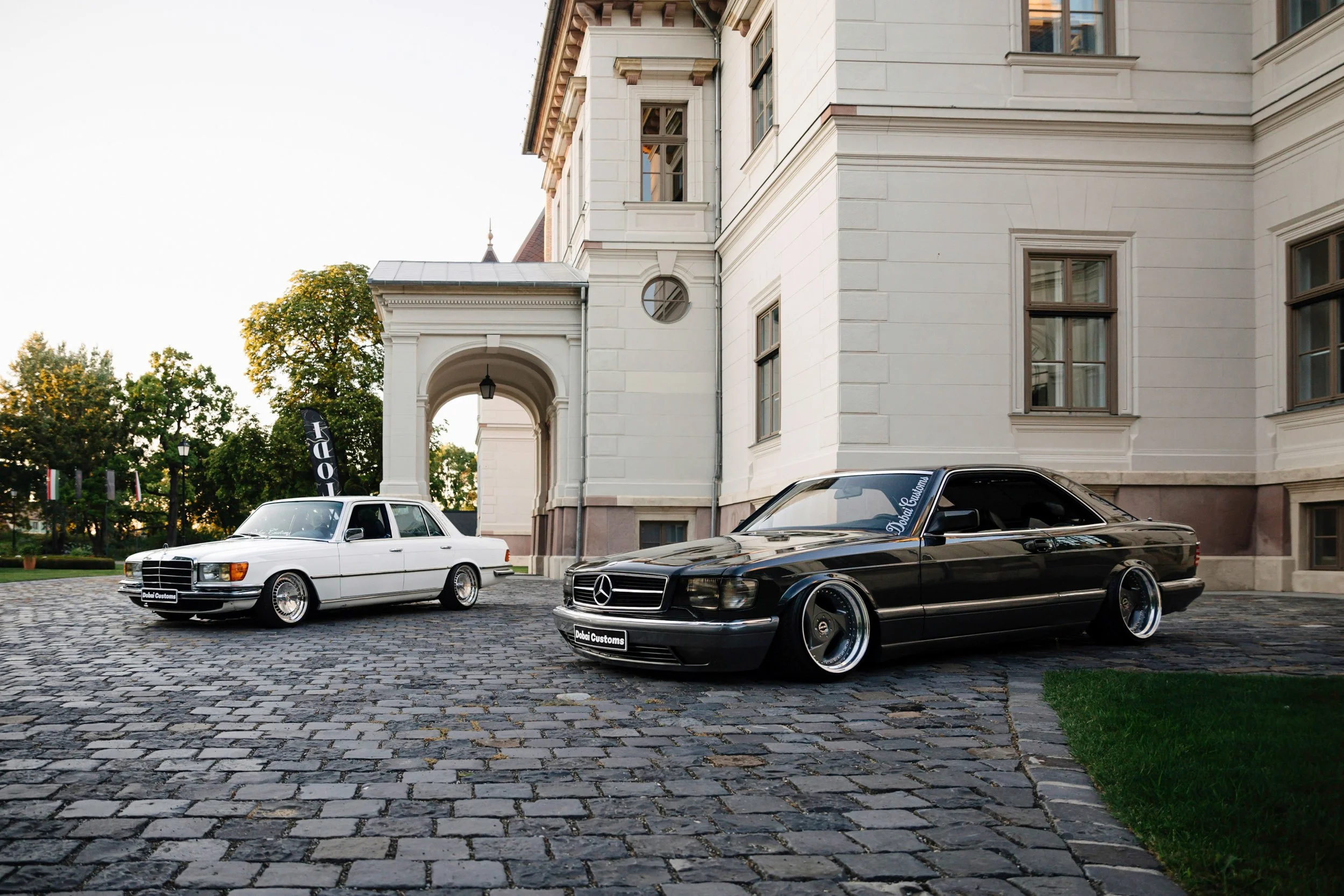 Two vintage Mercedes-Benz cars parked in front of a large, elegant white building with multiple windows. The cars are black and white, with custom badges and lowered suspensions. The cobblestone driveway adds to the historic and luxurious ambiance.