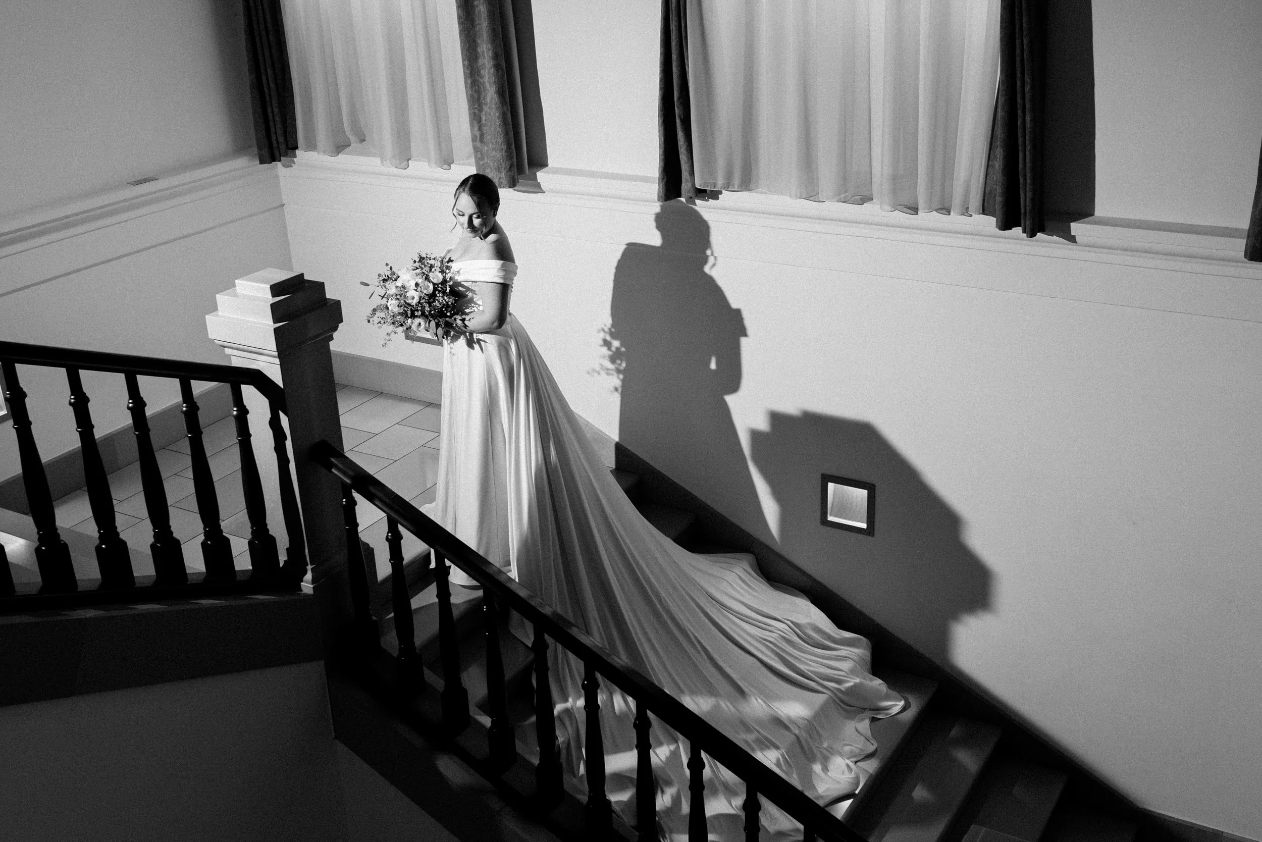 A bride in a strapless wedding gown holding a bouquet of flowers standing on a staircase with her long train flowing down the steps. Shadows cast on the wall behind her, with windows and curtains in the background.