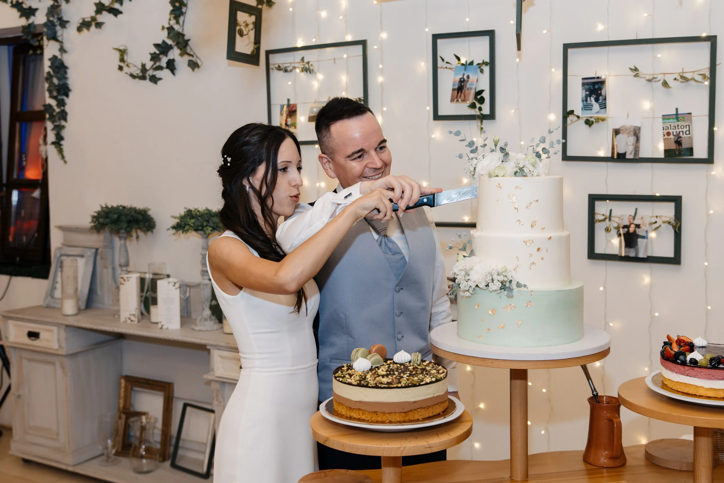 A woman in a white dress and a man in a vest are cutting a wedding cake together. The background is decorated with framed photos, greenery, and string lights. Several other cakes are on display on tables in the foreground.