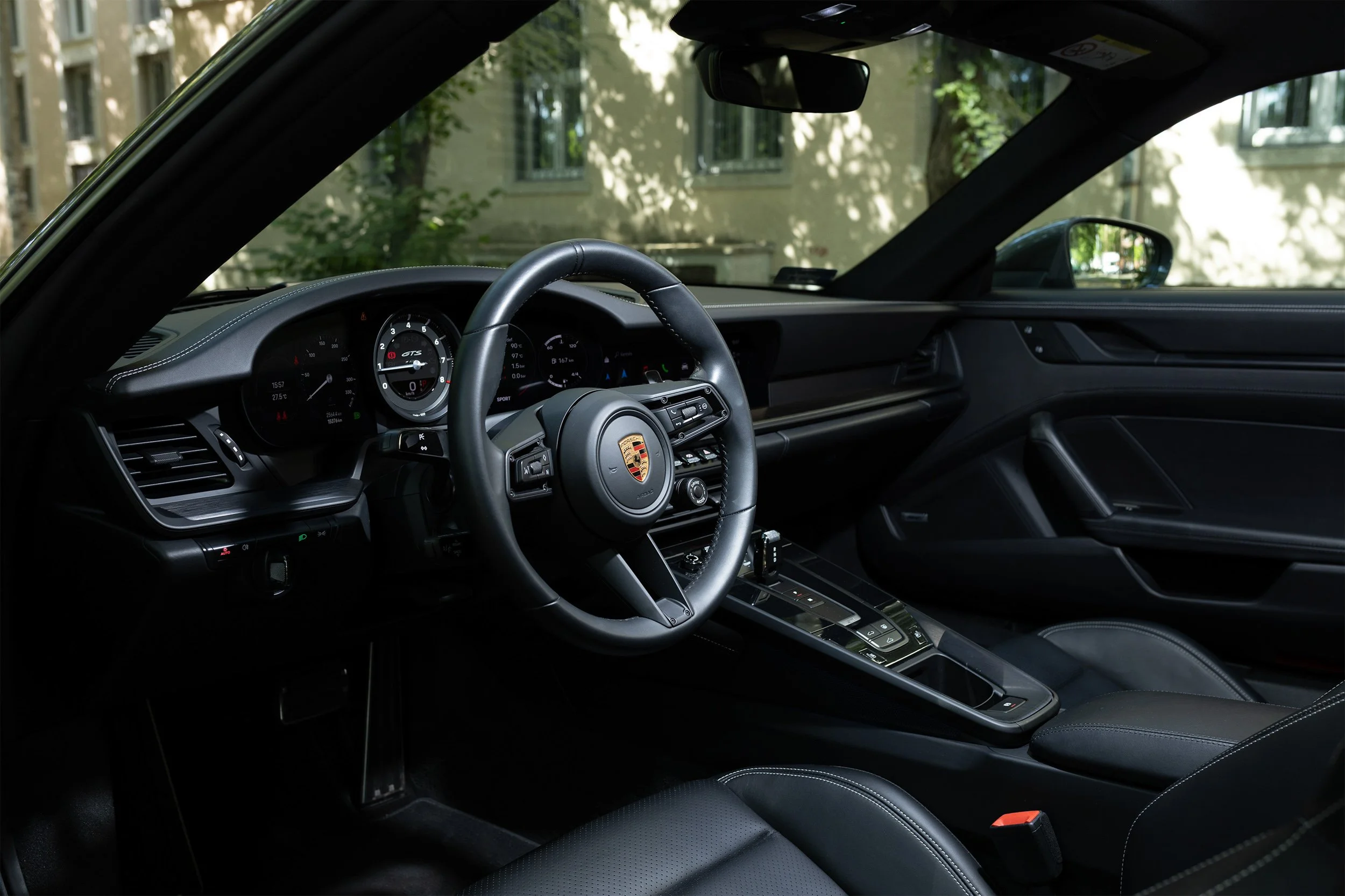 Interior of a Porsche sports car showing the dashboard, steering wheel with Porsche emblem, digital displays, and central console, with outdoor buildings and greenery visible through the windshield and side window.