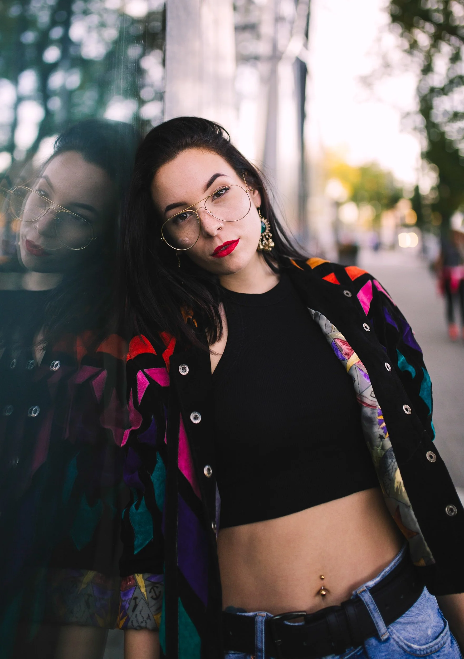 A woman with dark hair and freckles leaning her head against a reflective surface, wearing glasses, red lipstick, earrings, a colorful jacket, black crop top, and blue jeans with a belly piercing, outdoors with a blurred background of trees and stree