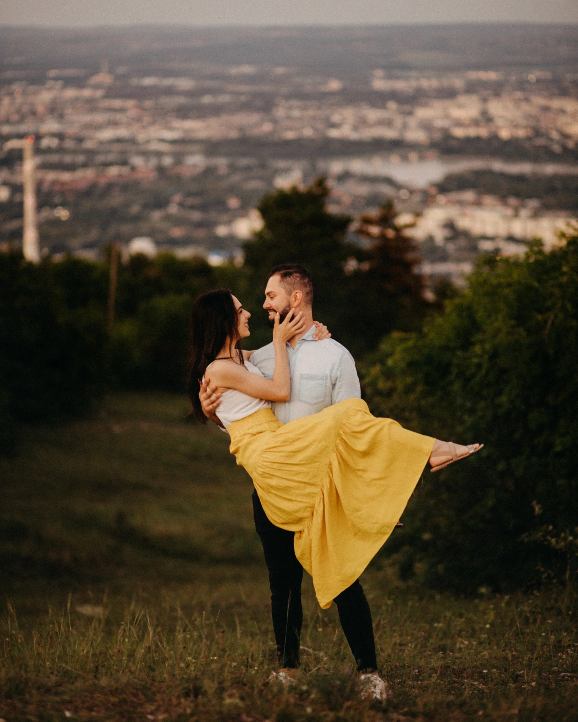 A couple dancing outdoors in a grassy field during sunset; the woman is wearing a yellow skirt and white top, and the man is in a white shirt and dark pants; they are smiling at each other with a cityscape and river in the background.