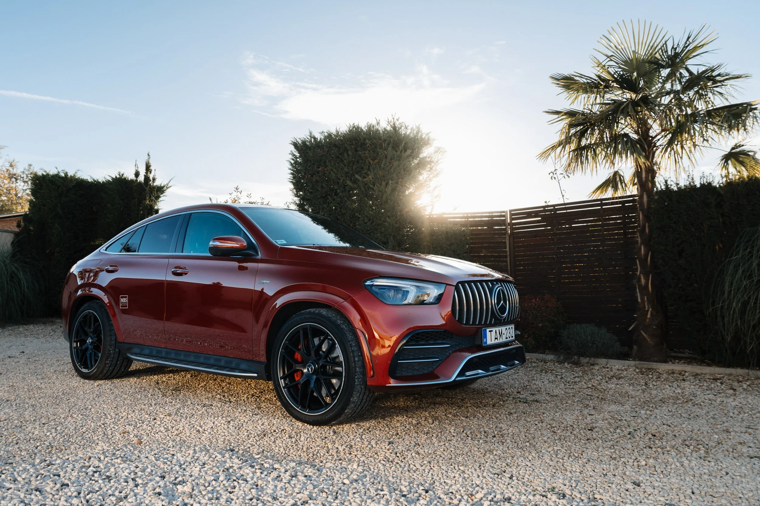 Red Mercedes-Benz SUV parked on a gravel driveway with a wooden fence and lush greenery, including palm trees, in the background during a bright sunny day.
