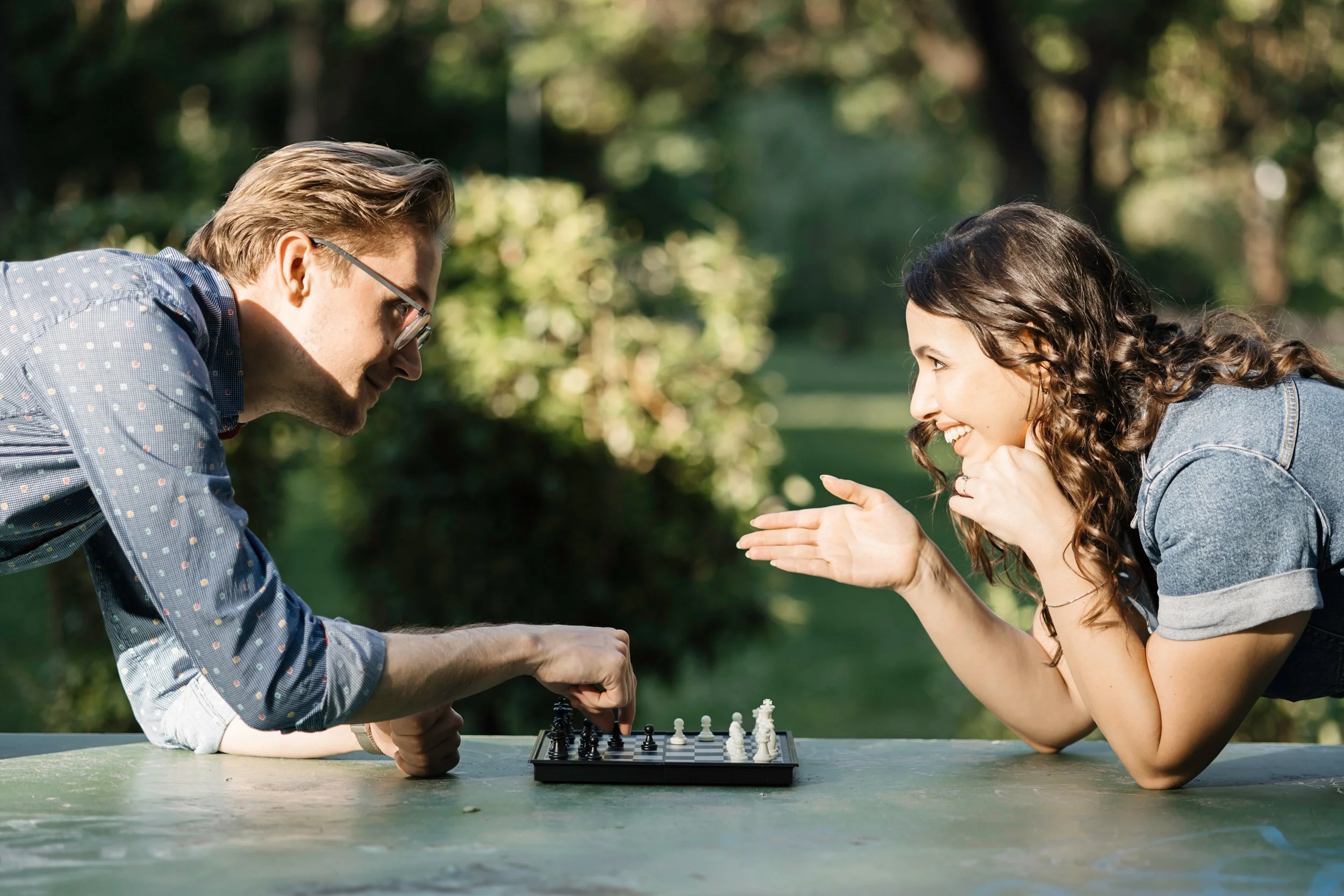A man and a woman are playing chess outdoors at a table surrounded by trees, engaging and smiling at each other.