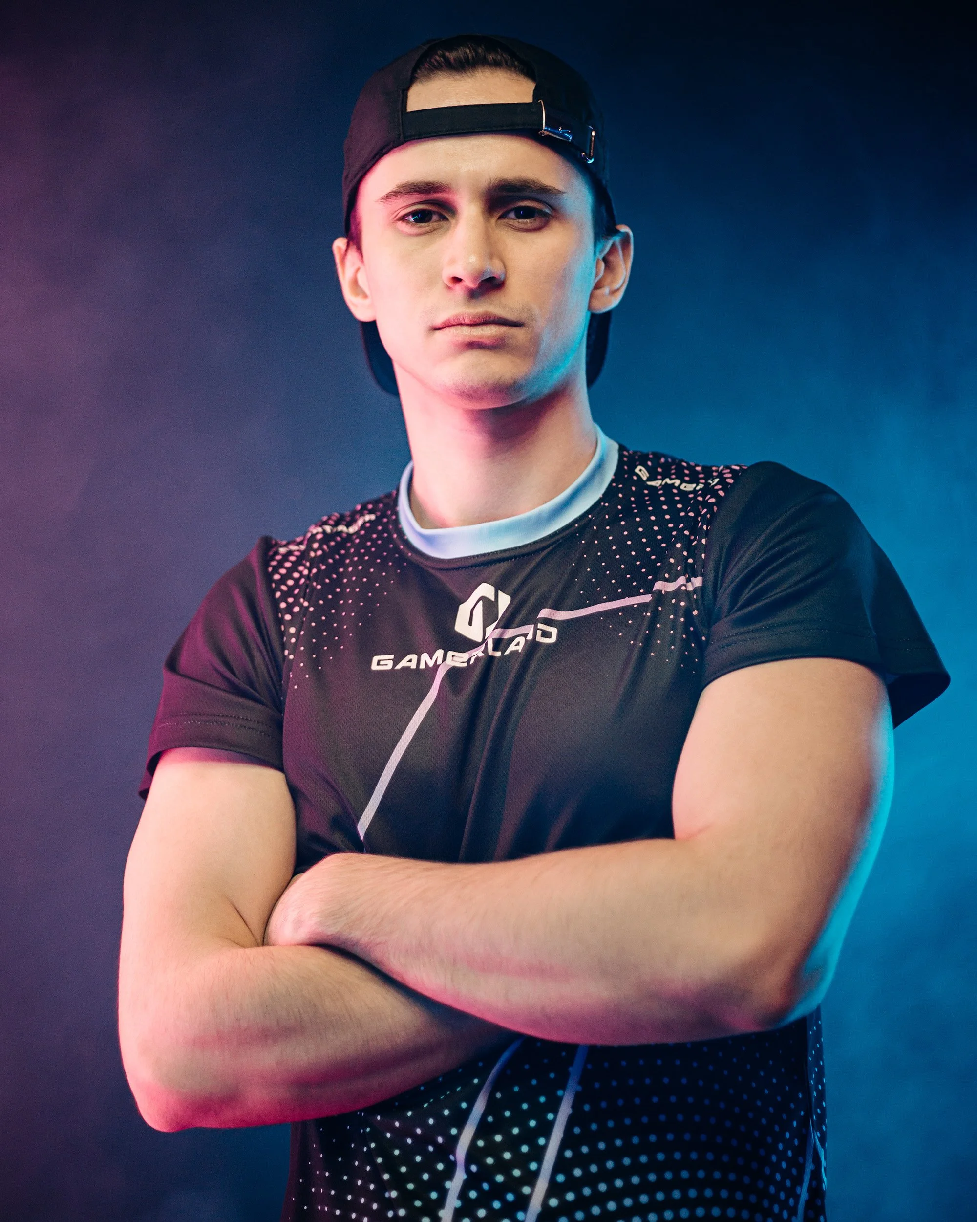 Young man wearing a black baseball cap backwards and a black gaming jersey with white dots and a logo, crossing his arms, with a serious expression, in a colorful, neon-lit studio background.