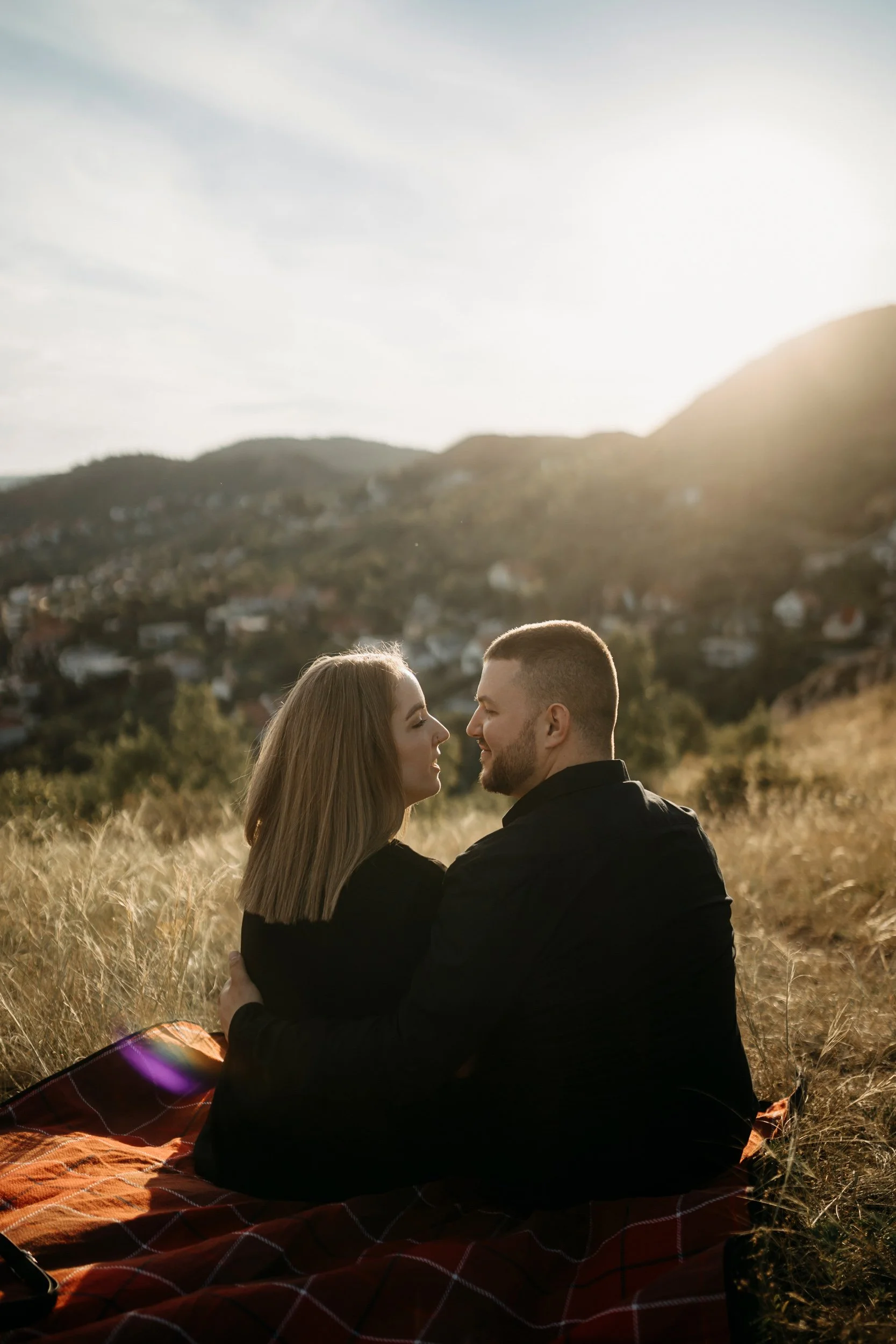 A couple sitting closely in a field during sunset, looking into each other's eyes with mountains in the background.