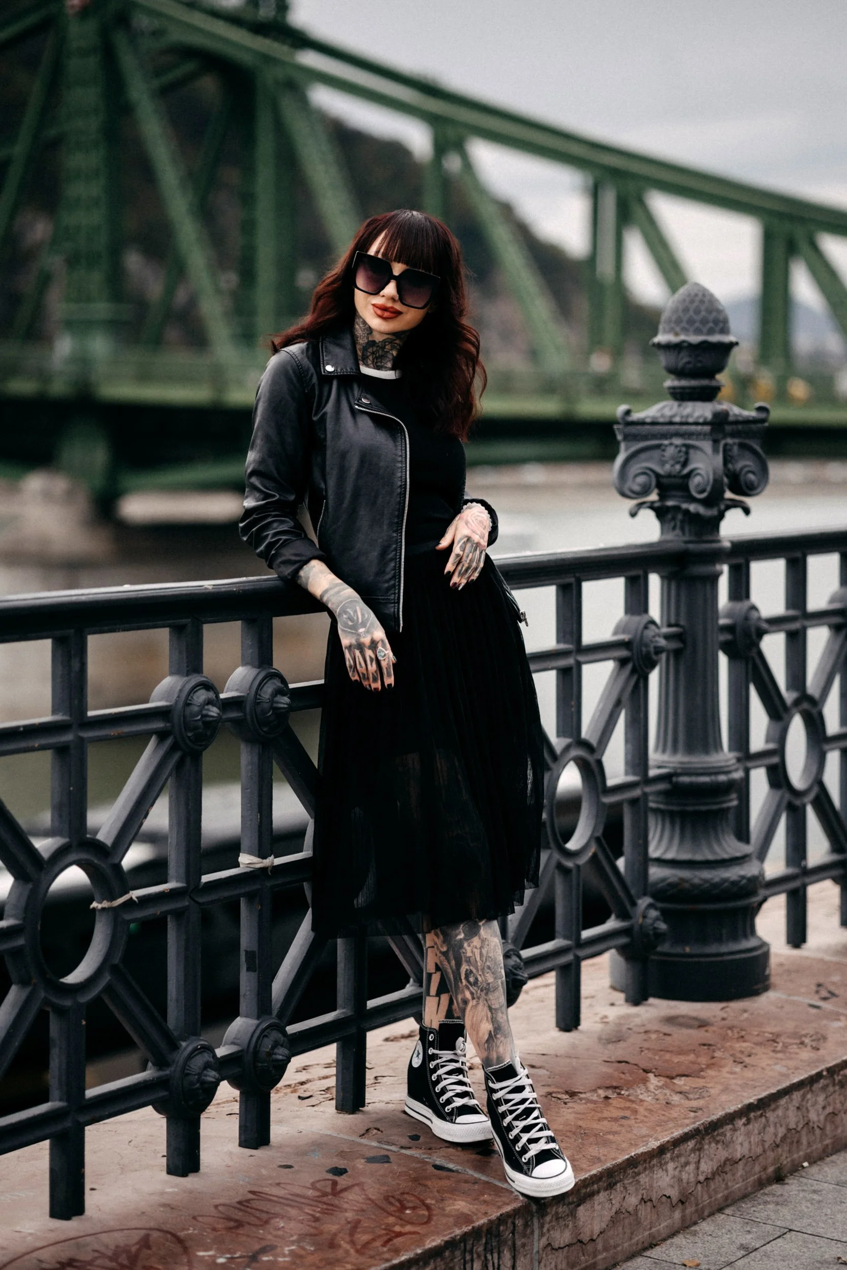 A woman with tattoos, dark lipstick, and sunglasses wearing a leather jacket and black dress standing by a decorative black iron railing near a river with a green bridge in the background.