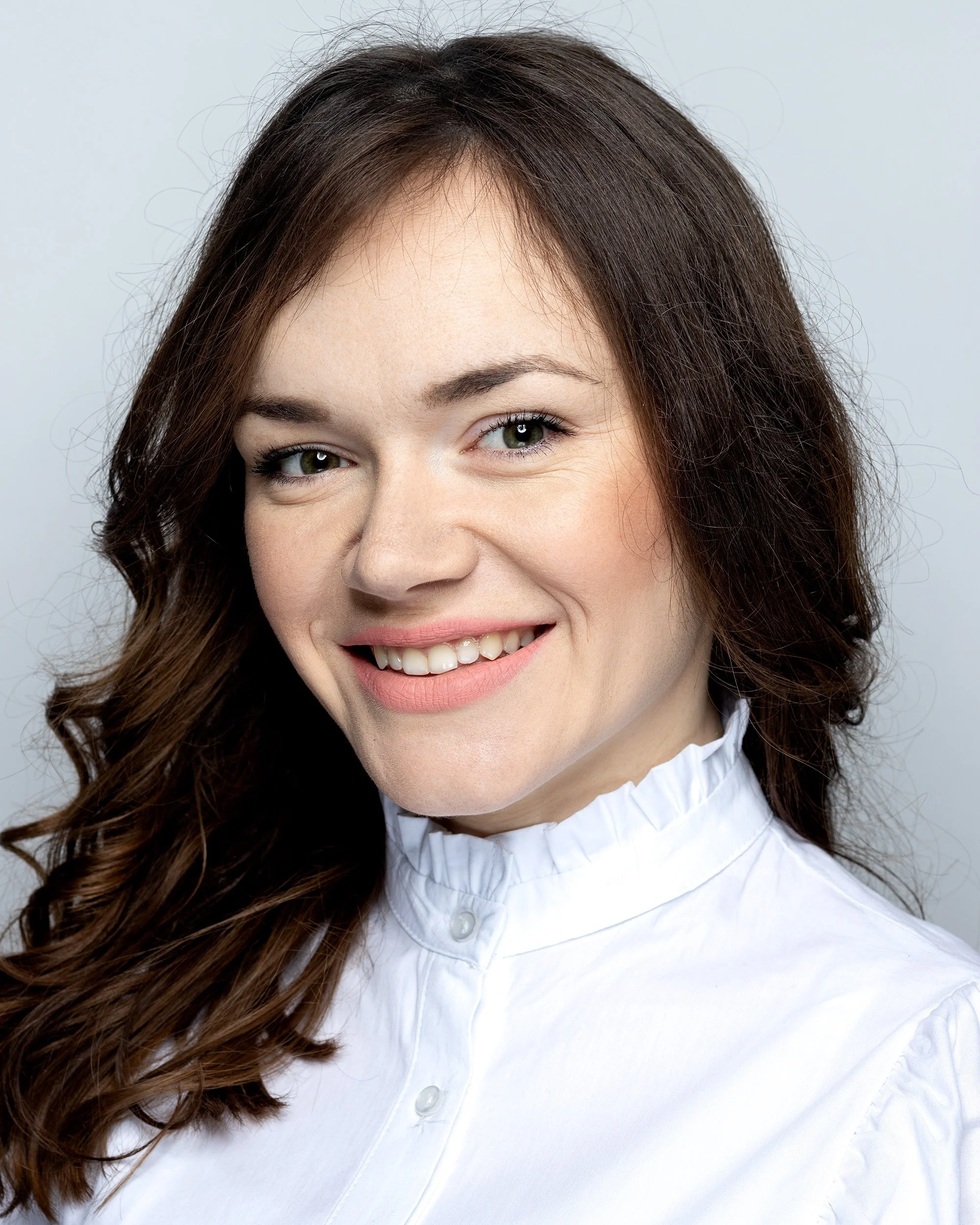 Close-up of a woman with long brown hair, smiling, wearing a white collared shirt.