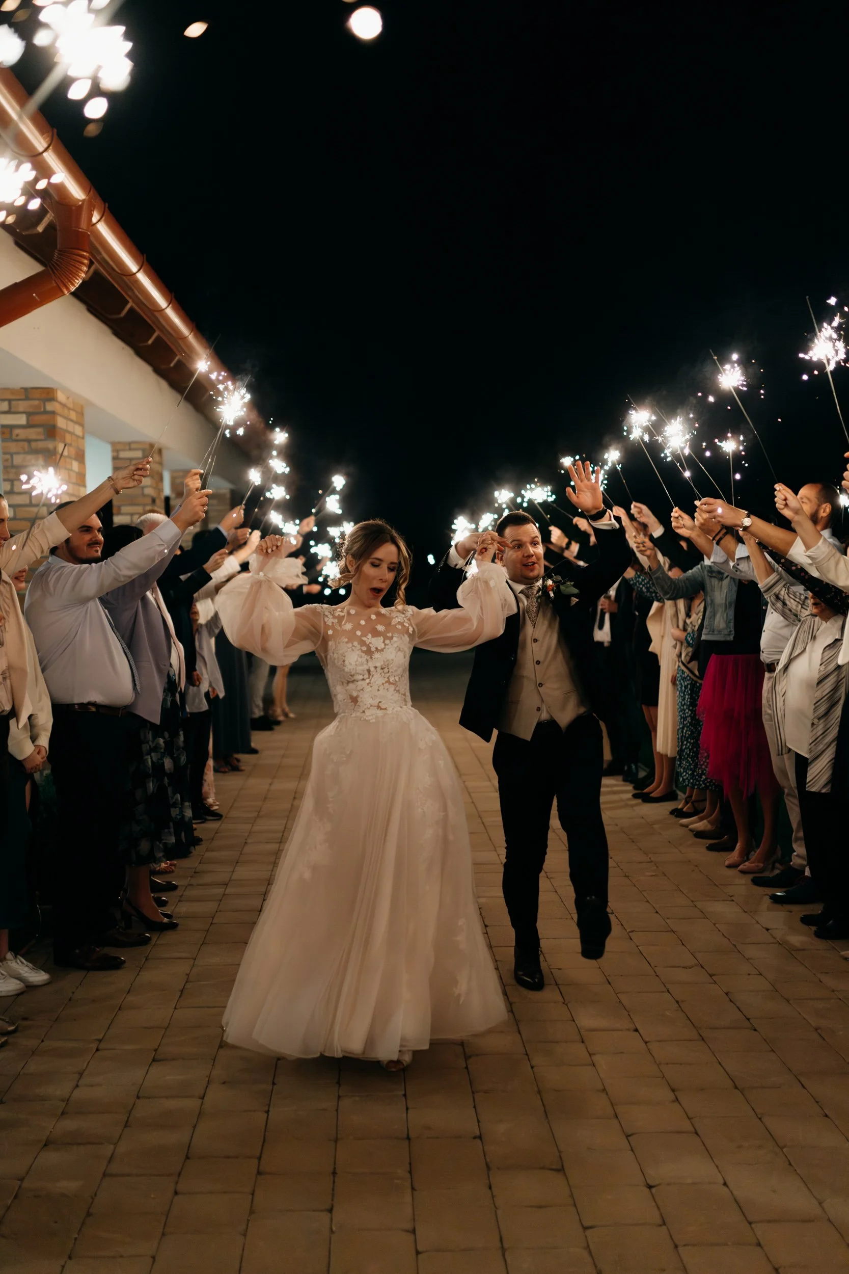 A bride and groom walking together through a line of guests holding sparklers during a celebration at night.