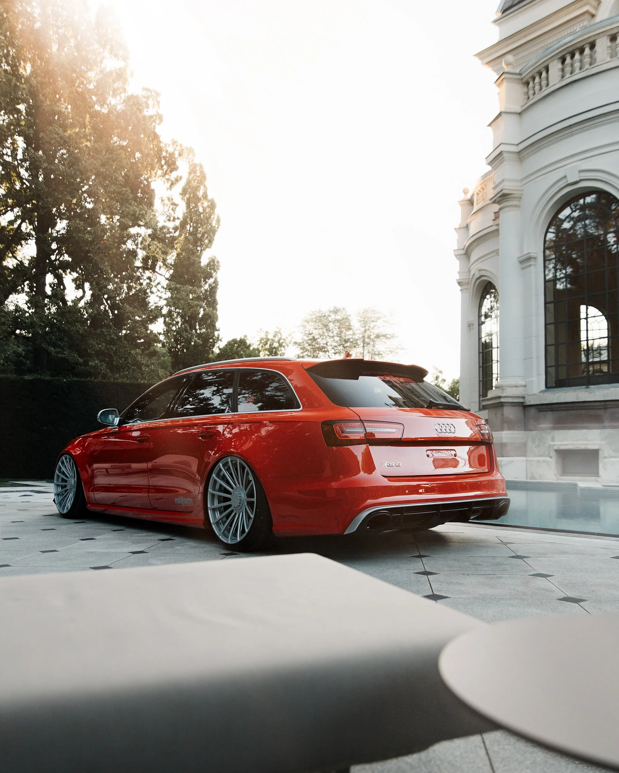 A red Audi RS6 car parked outside a white building with tall windows and ornate architectural details, with trees and sunlight in the background.