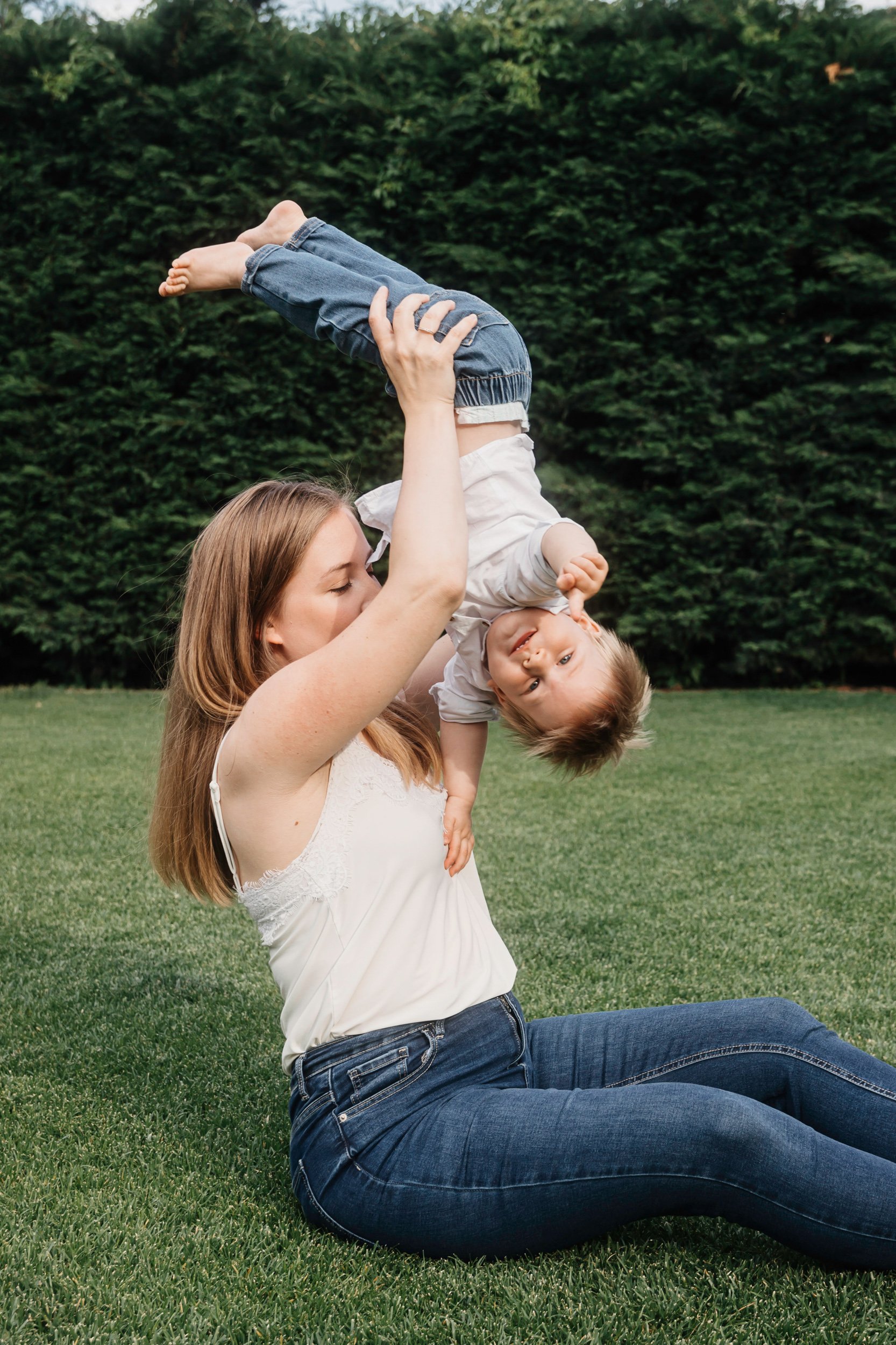 A woman sitting on grass and lifting a young boy in the air, both smiling, with a background of trees.