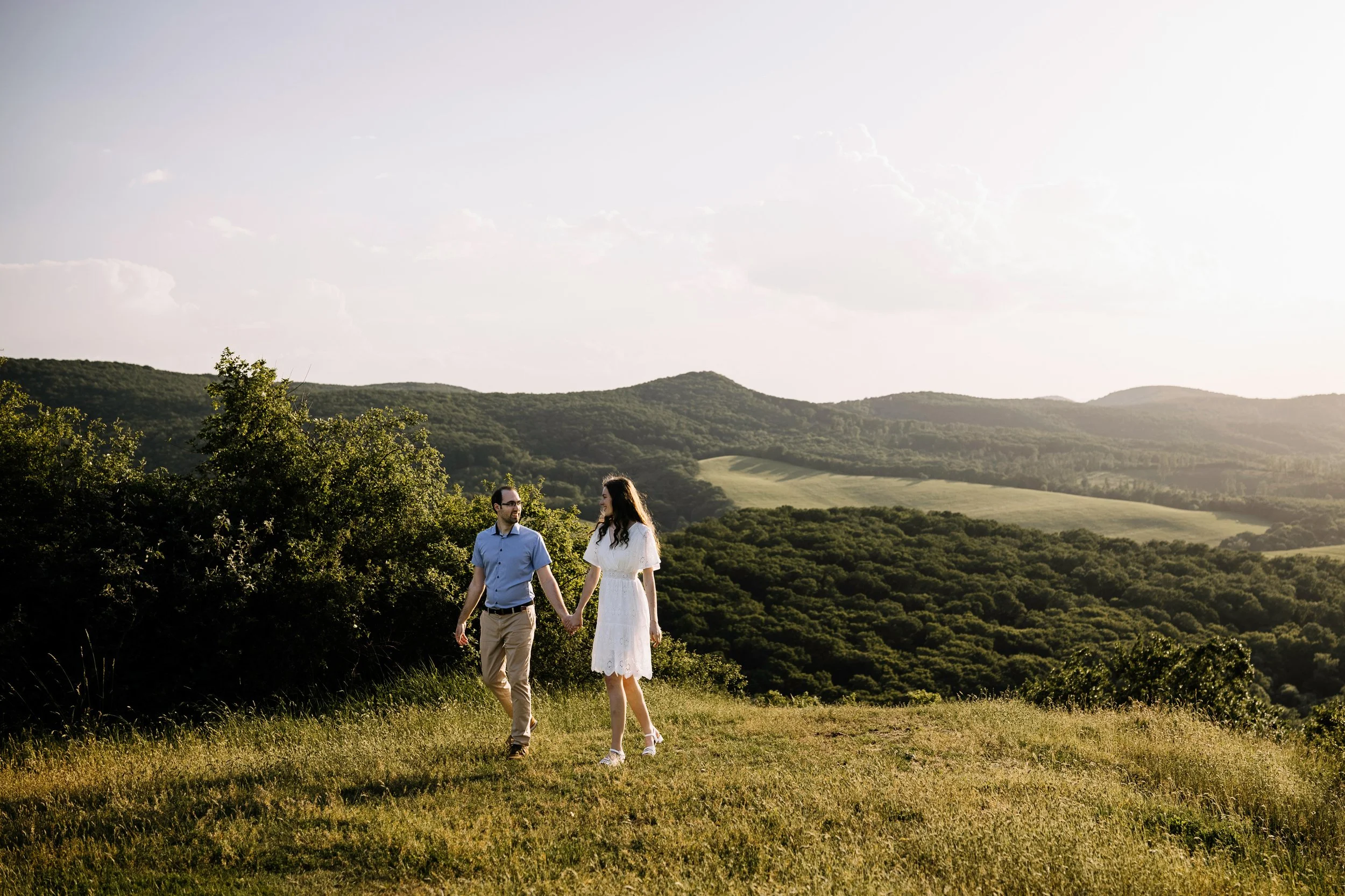 A couple holding hands and walking through a grassy field with rolling hills and trees in the background, under a clear sky.