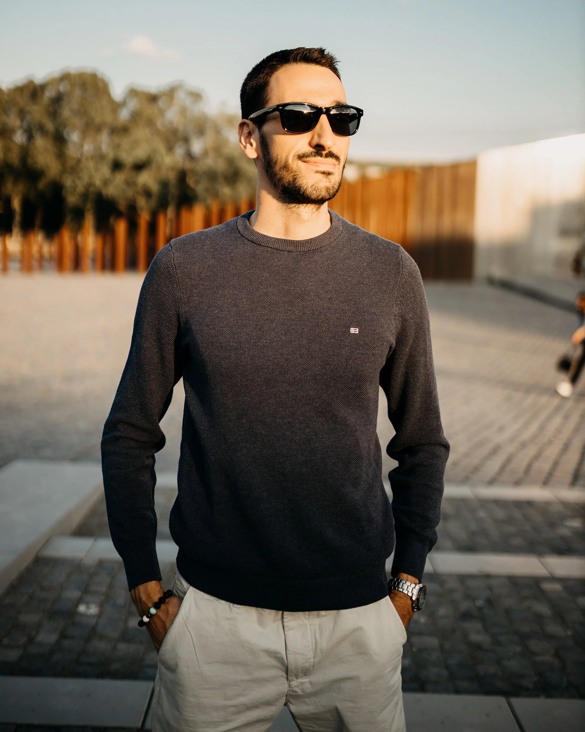 A man with a beard and dark hair wearing sunglasses, a grey sweater, and beige pants, stands outdoors on a cobblestone path with a wooden fence and trees in the background, during sunset.
