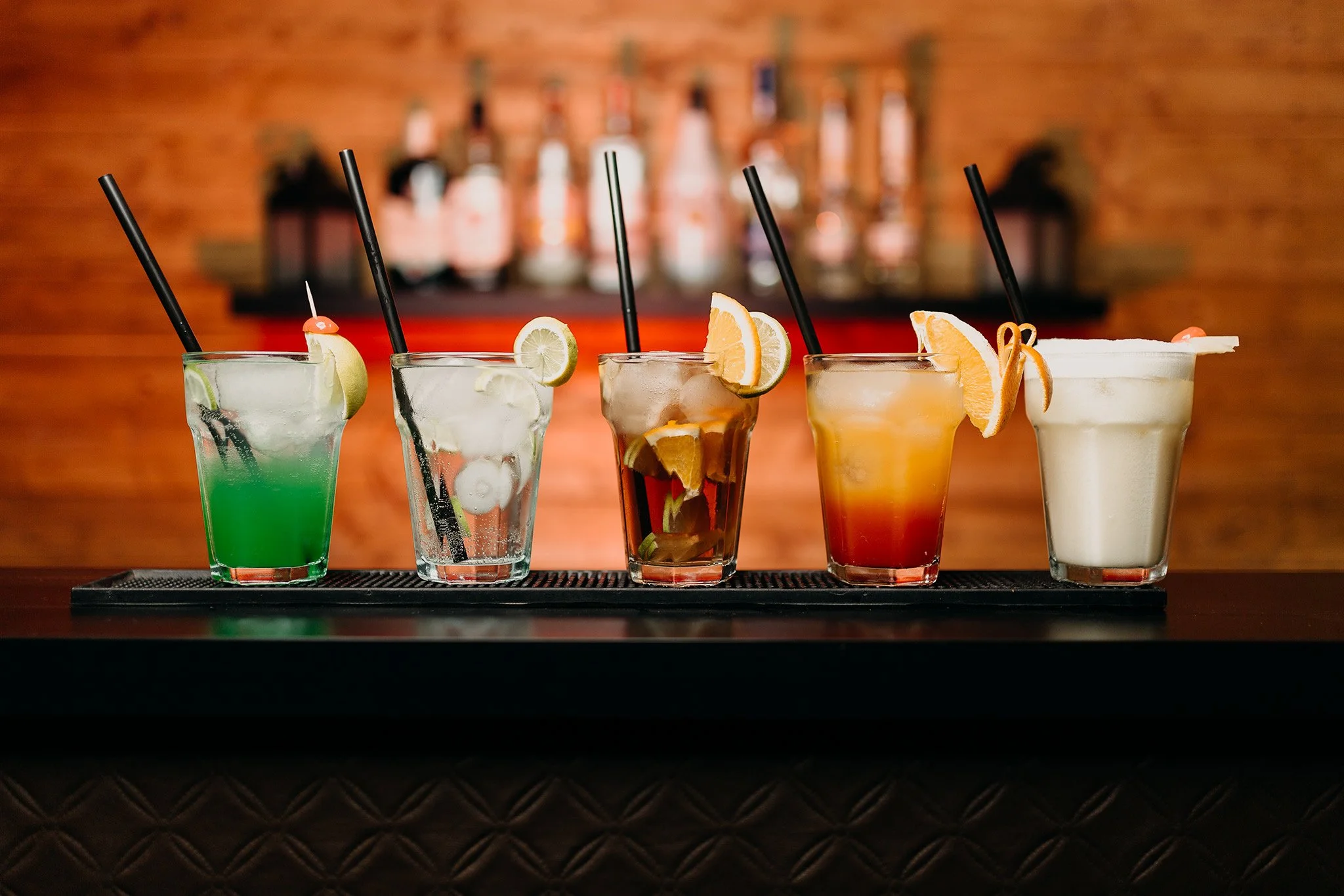A row of five colorful cocktails on a black bar mat, with a background of warm wooden wall and bottles of alcohol. The cocktails have garnishes of lemon, lime, orange slices, and cherries.