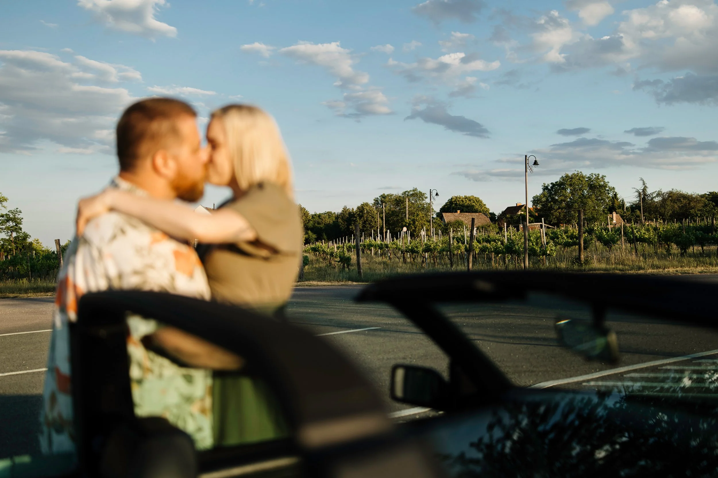 A couple sharing a kiss outdoors with vineyard and houses in the background, seen from inside a car.